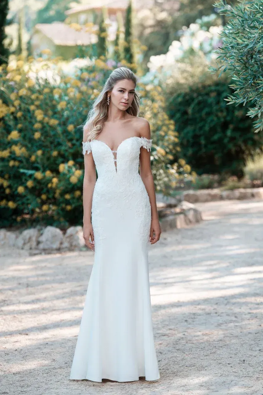 Woman in off-shoulder white wedding dress, standing on a path outdoors, looking towards the camera.