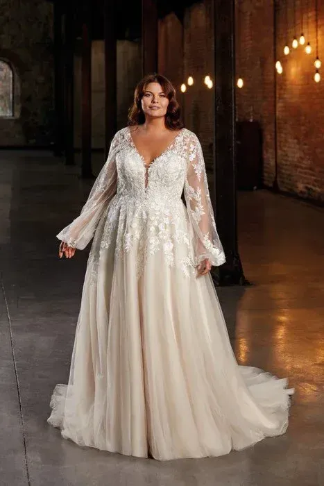 Woman in a white wedding dress with long lace sleeves and a plunging neckline, standing in an industrial setting.
