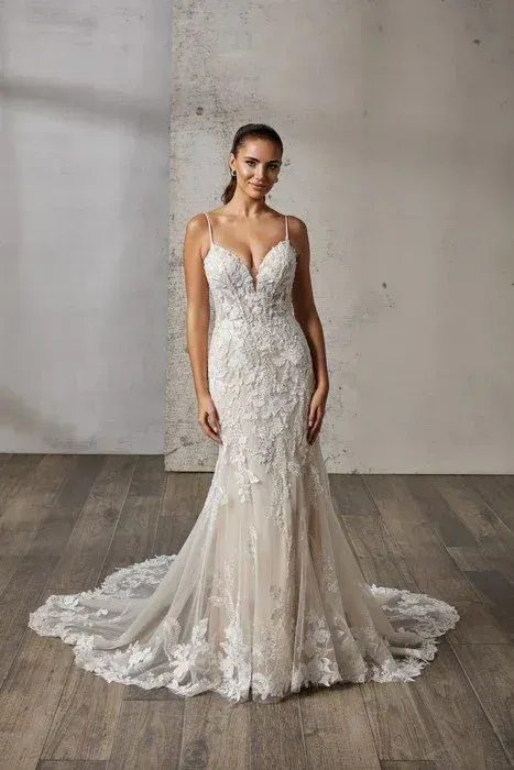 Woman in a white, form-fitting wedding dress with a long train, standing on a wooden floor.