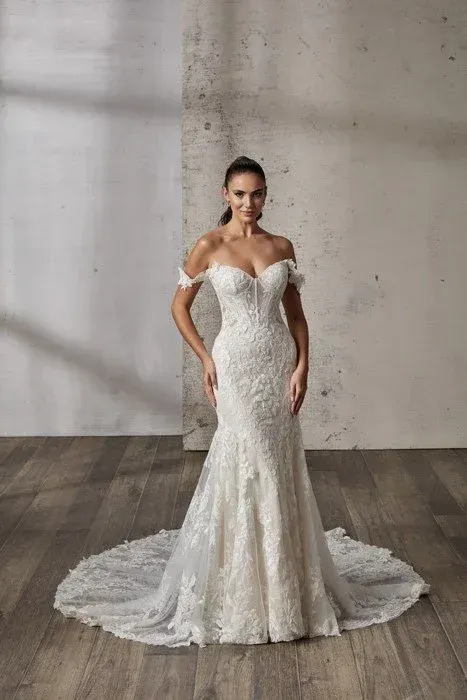 Woman in off-shoulder lace wedding dress with mermaid skirt, standing in a studio with a wood floor.