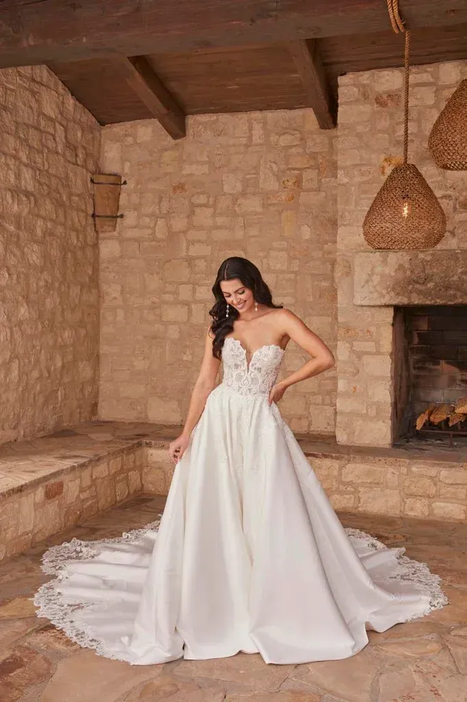 Bride in strapless white gown with train, standing in front of stone fireplace.