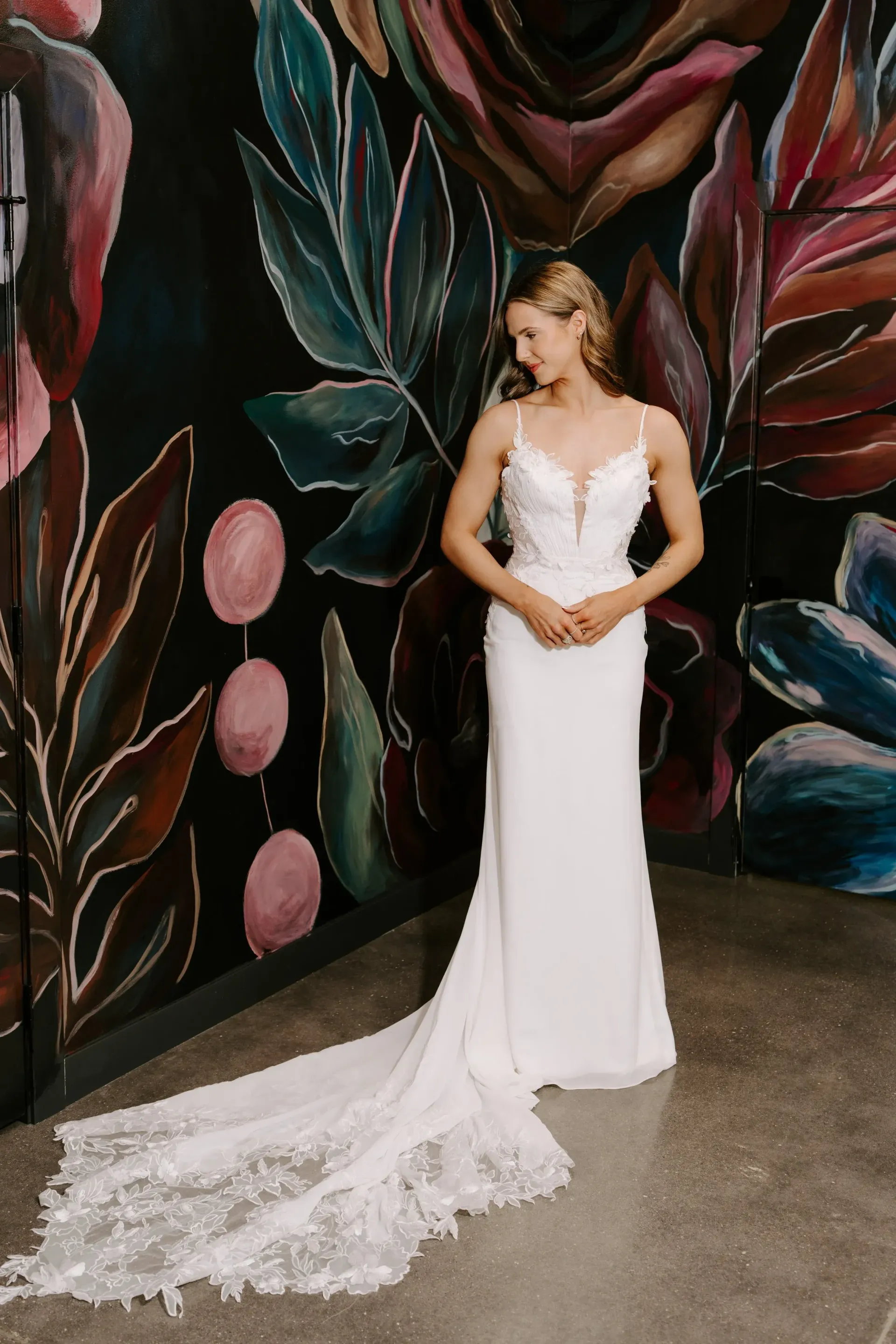 Woman in a white wedding dress with a long train, standing against a colorful mural.