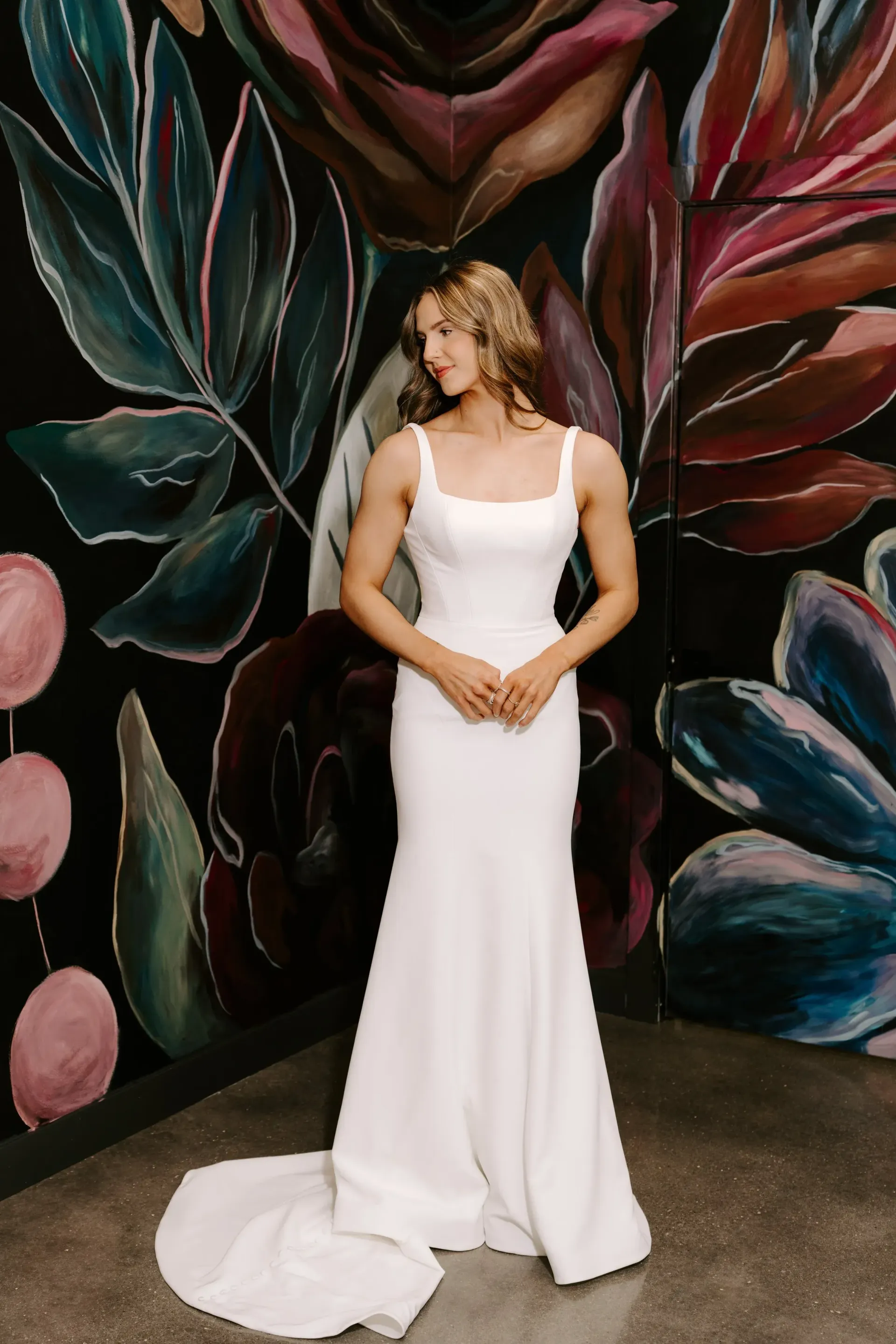 Woman in white wedding dress poses against floral mural.