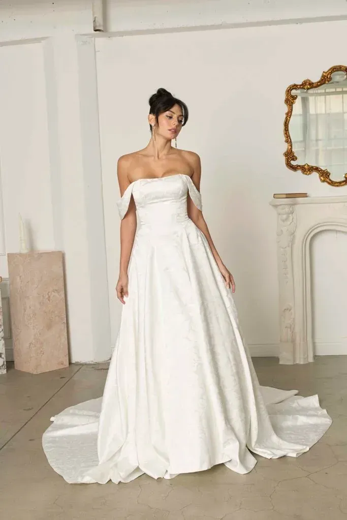 Woman in white off-the-shoulder wedding dress, posing in a bright room.
