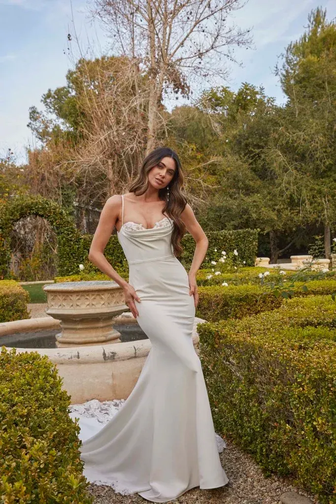 Woman in a white satin wedding dress with a cowl neckline and mermaid skirt poses in a garden.