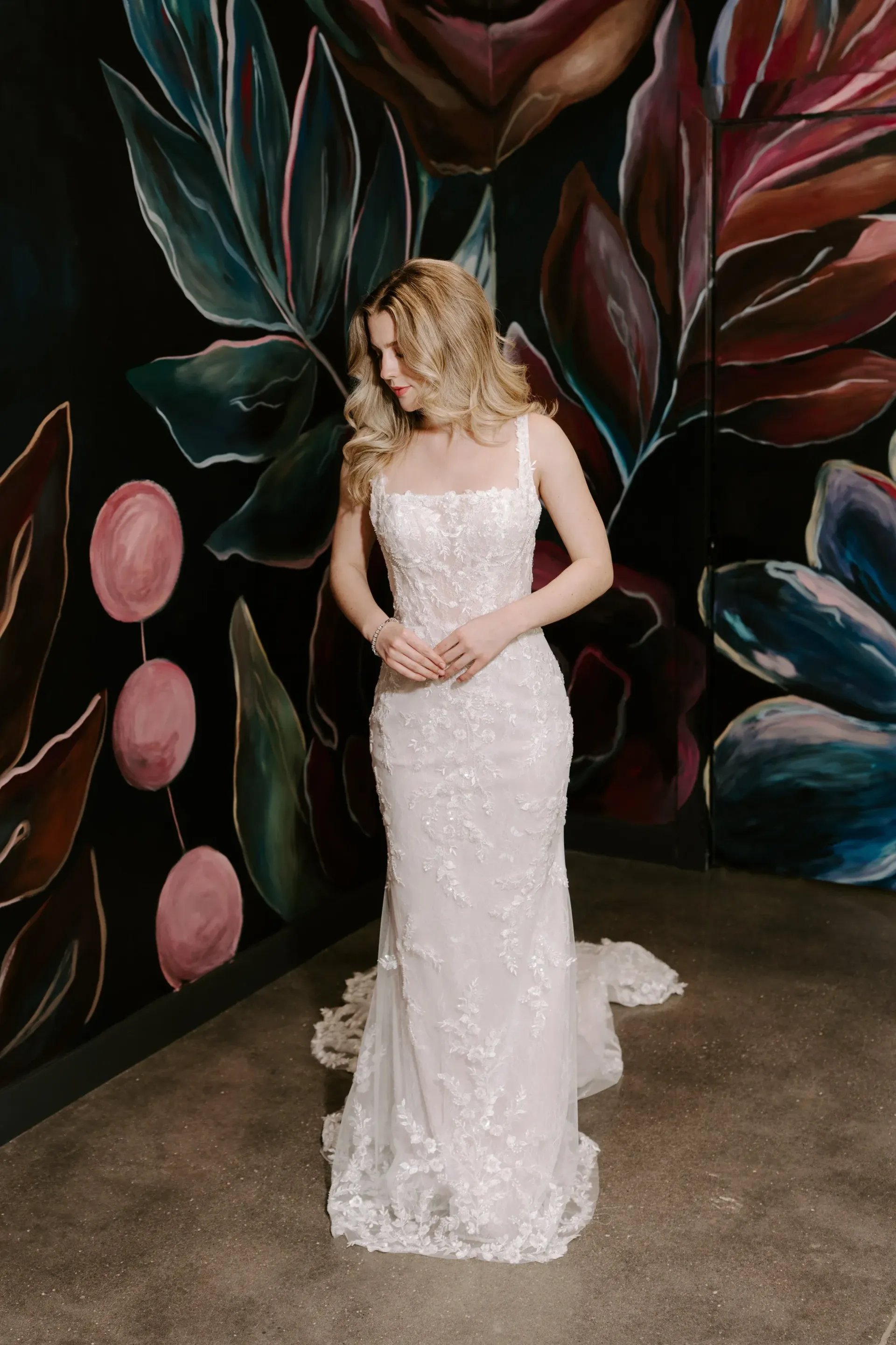 Blonde woman in a white wedding gown poses by a floral mural.