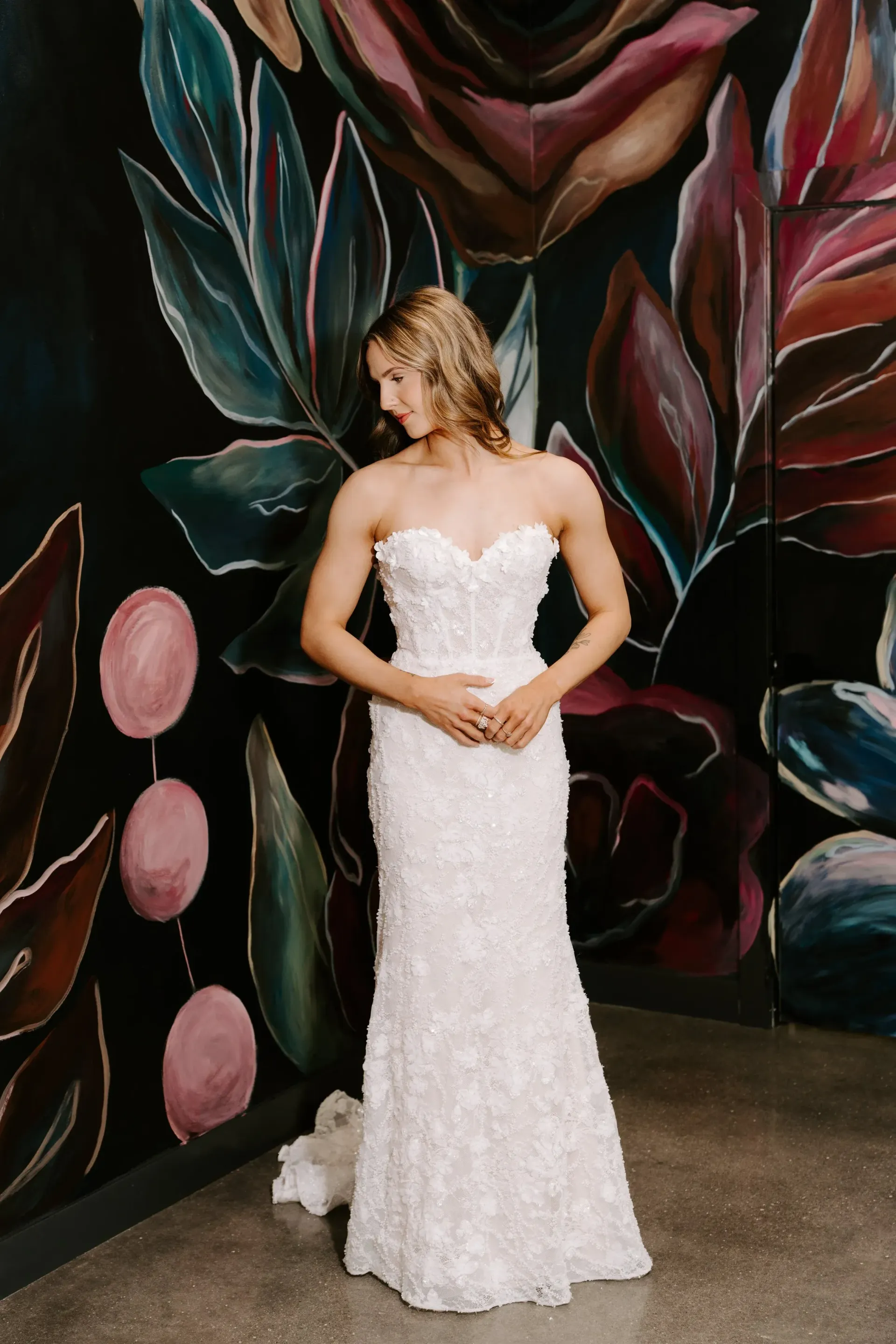 Woman in a white floral wedding dress posing in front of a colorful floral mural.