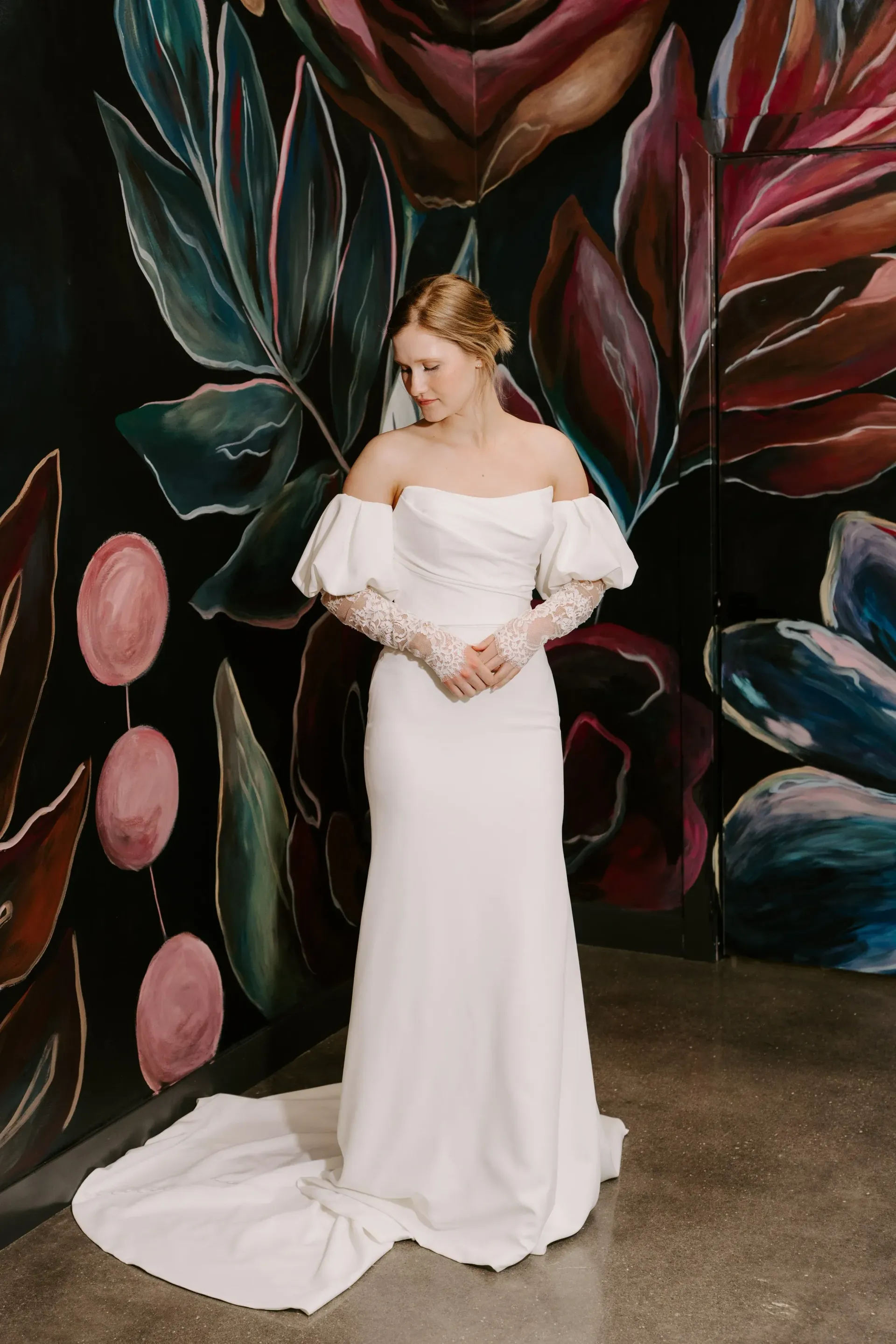 Bride in white dress with lace gloves, standing against a floral mural.