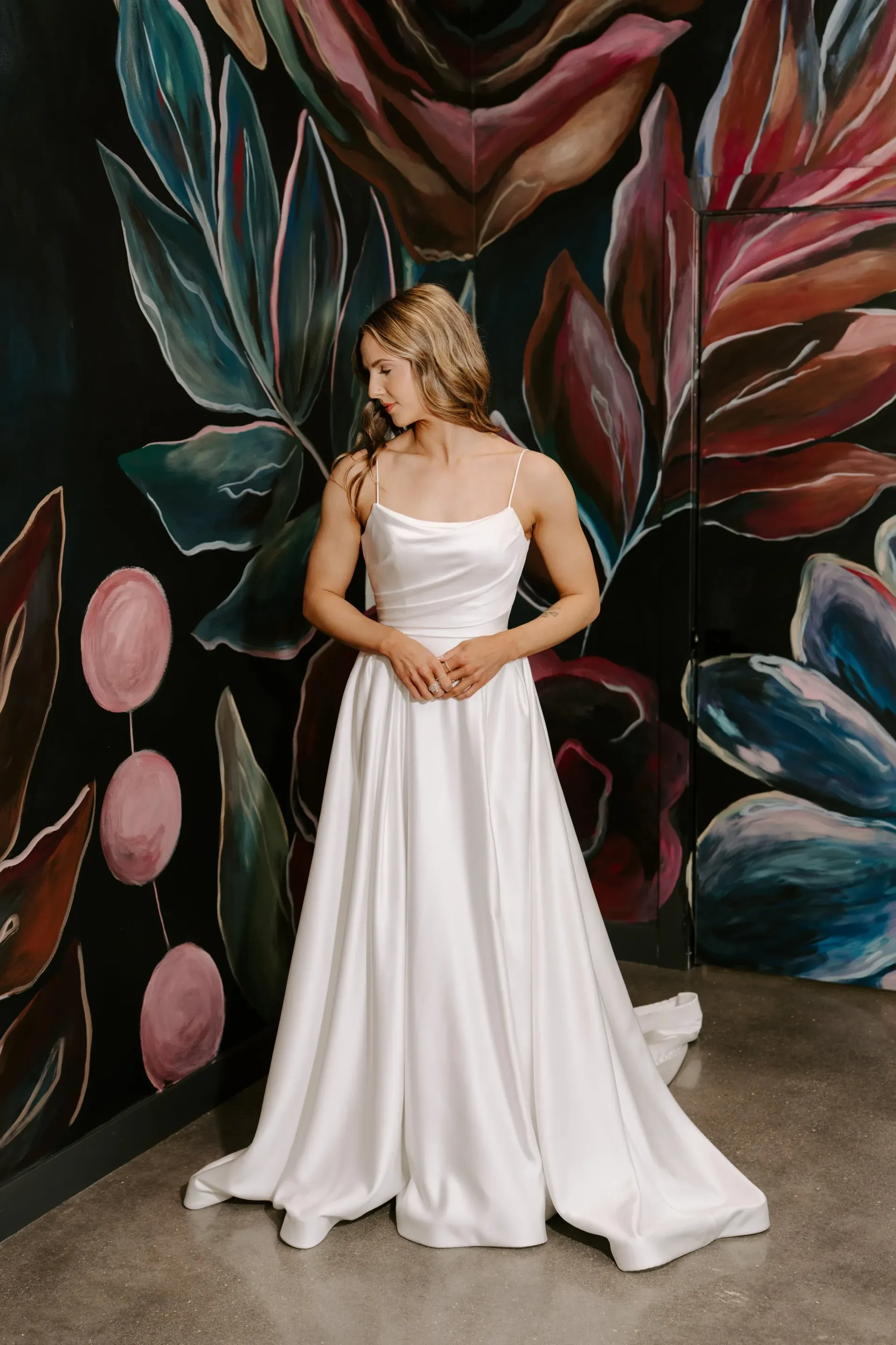 Woman in white satin dress poses in front of a floral mural.