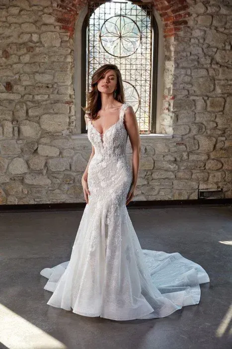 Woman in a white, embellished mermaid wedding dress with a long train, standing in a stone room.