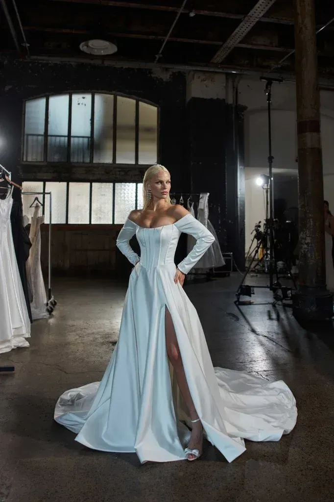 Blonde woman in white wedding dress with high slit, hands on hips, in a studio.