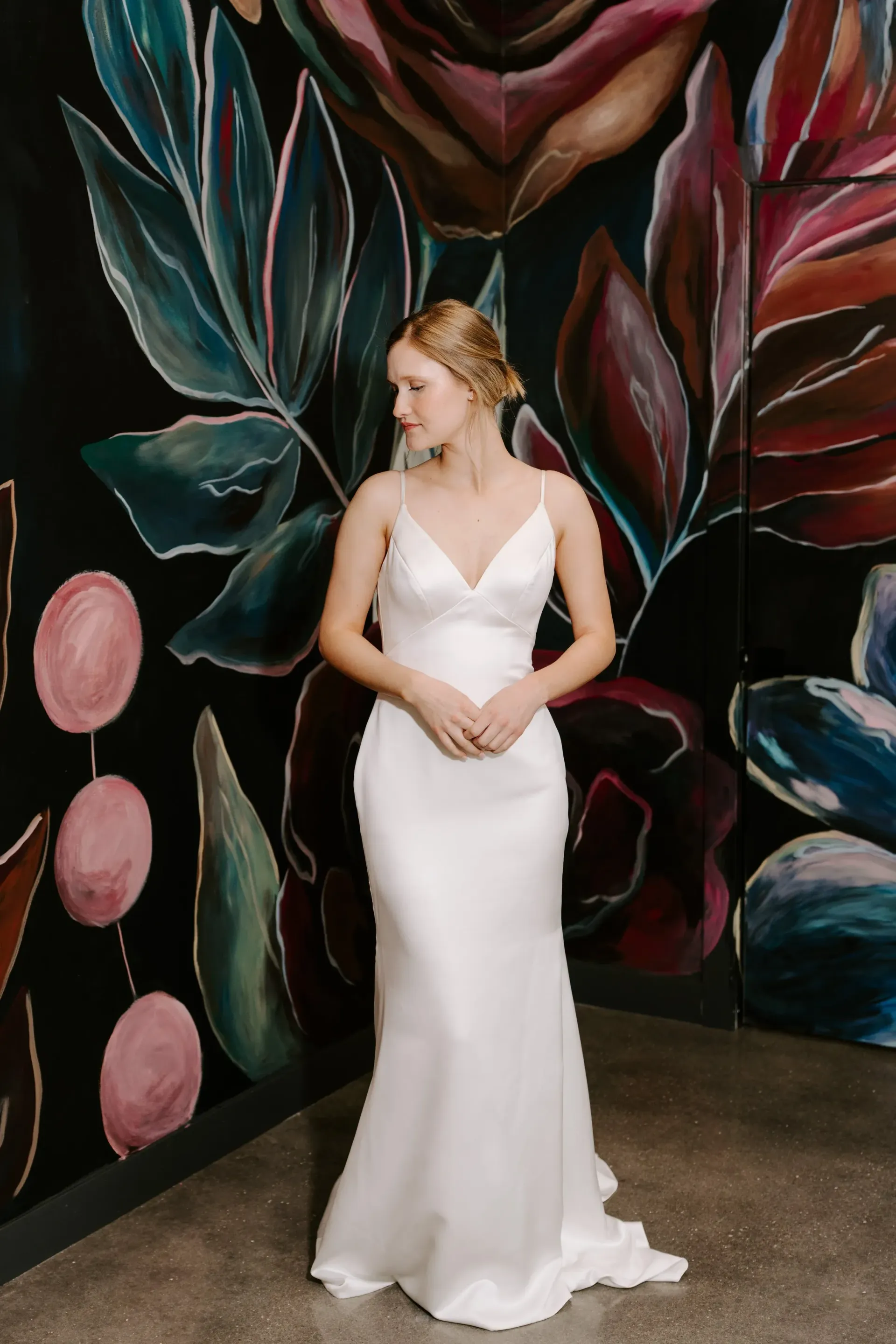 Woman in a white wedding dress stands before a floral painted wall, looking sideways.