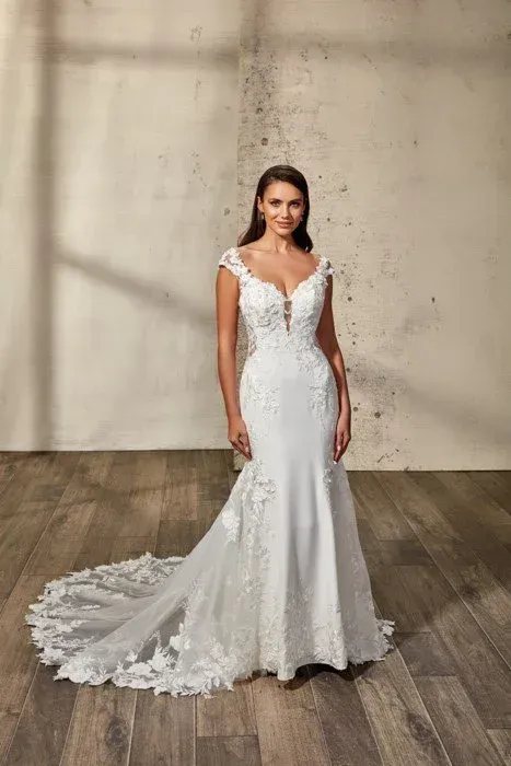 Woman in a white mermaid-style wedding dress with floral lace details, standing in a rustic setting.