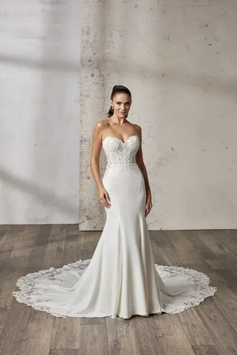 Woman in a white strapless mermaid wedding dress with lace detail on bodice and train, standing on a wood floor.