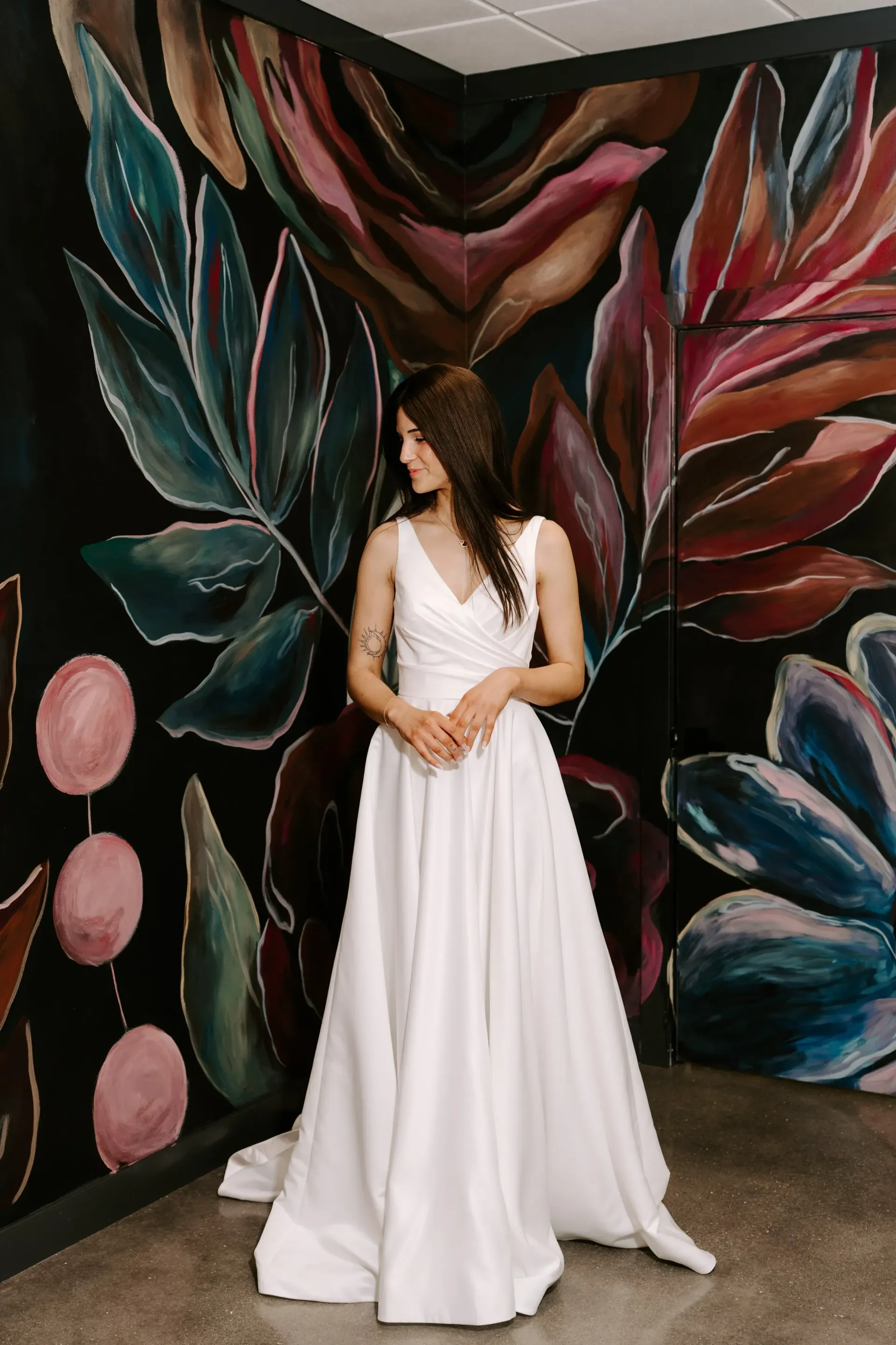Woman in white gown poses against a black wall with colorful painted flowers.
