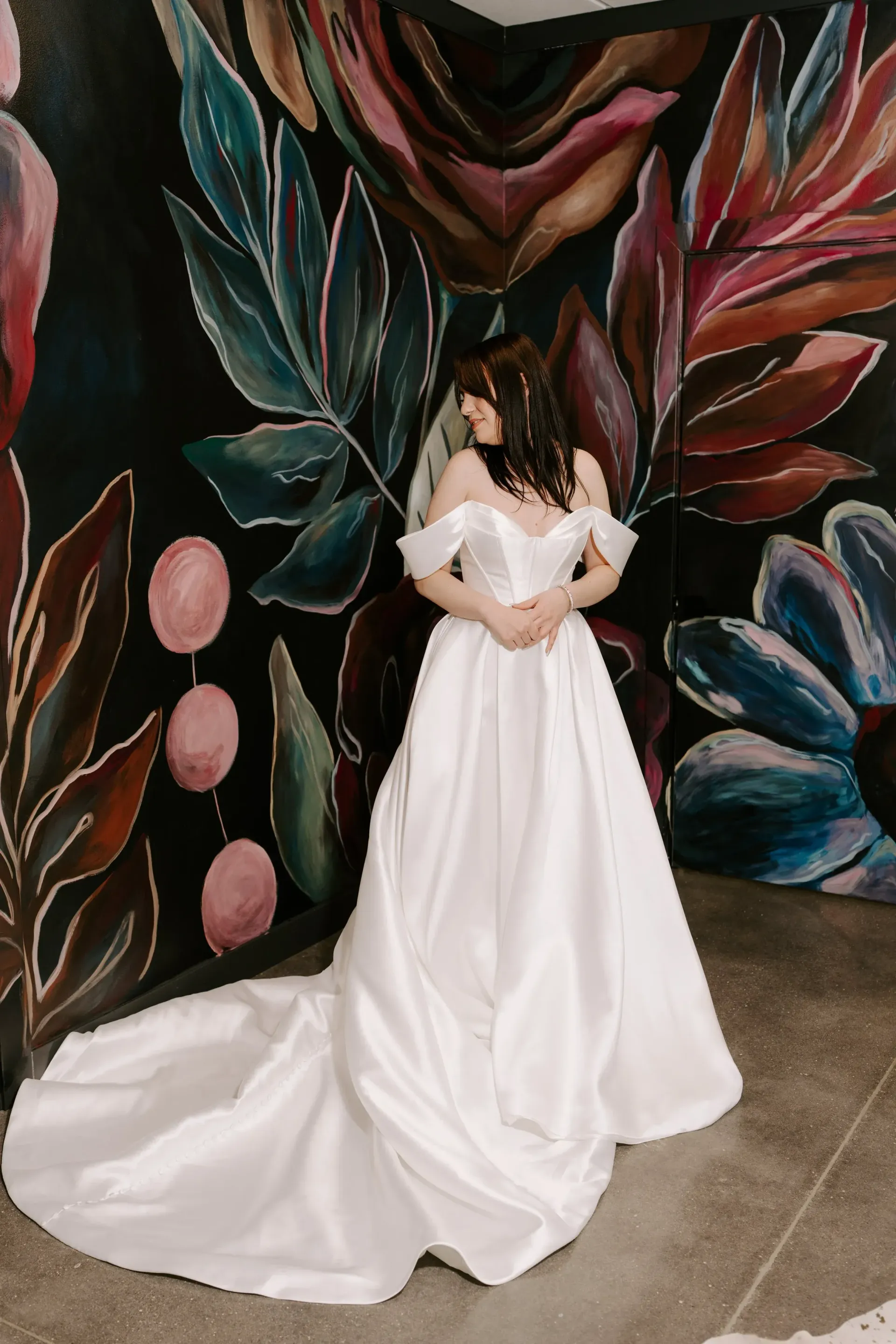 Woman in white off-shoulder wedding dress, train flowing, standing before a floral painted wall.
