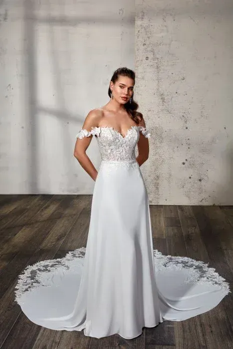 Woman in off-the-shoulder white wedding dress with long train, standing in front of a textured wall.
