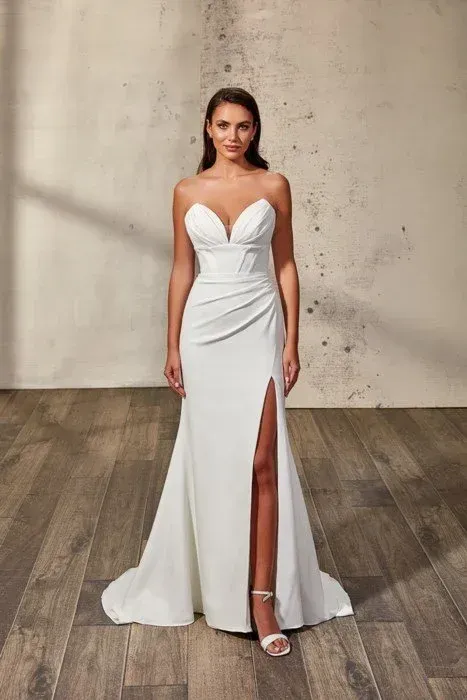 Woman in a white strapless wedding dress with a thigh-high slit, standing in front of a wall and wooden floor.