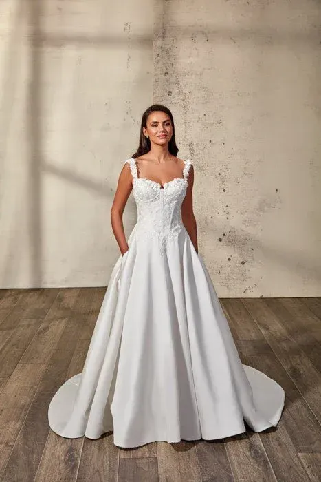 Woman in a white wedding dress with a beaded bodice and full skirt, standing indoors.