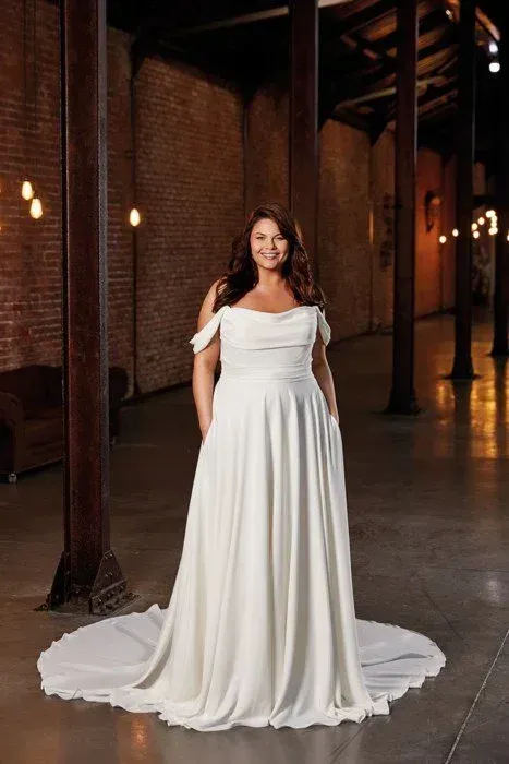 Woman in a white off-the-shoulder wedding dress stands in a brick-walled venue, smiling with hands in pockets.