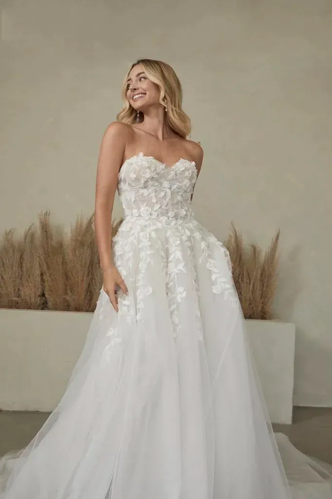 Blonde woman in white strapless wedding dress smiles, standing indoors.