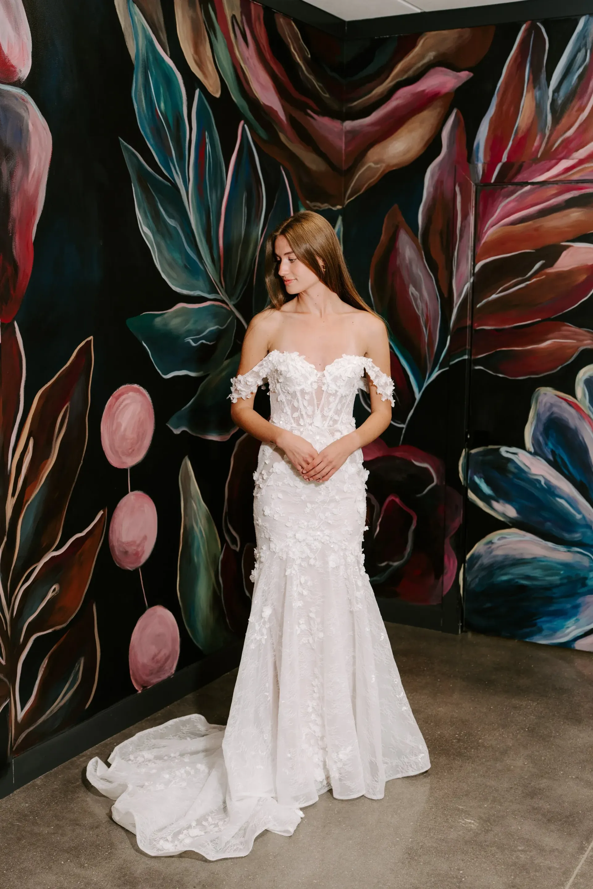 Woman in white lace wedding dress stands by floral mural.