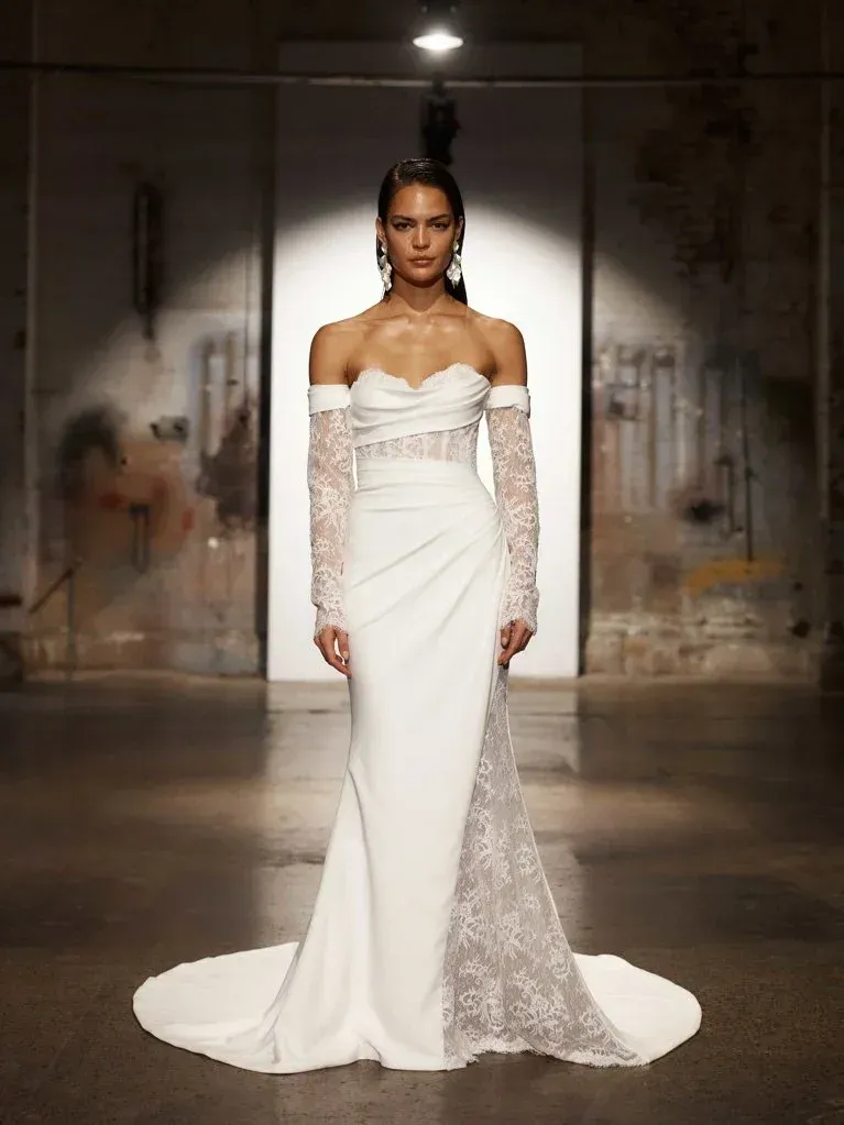 Woman in white wedding gown with lace sleeves and side panel, standing in a dim industrial space.