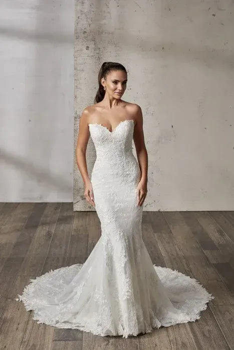 Bride in a strapless, lace mermaid wedding dress with a long train; wooden floor, neutral backdrop.