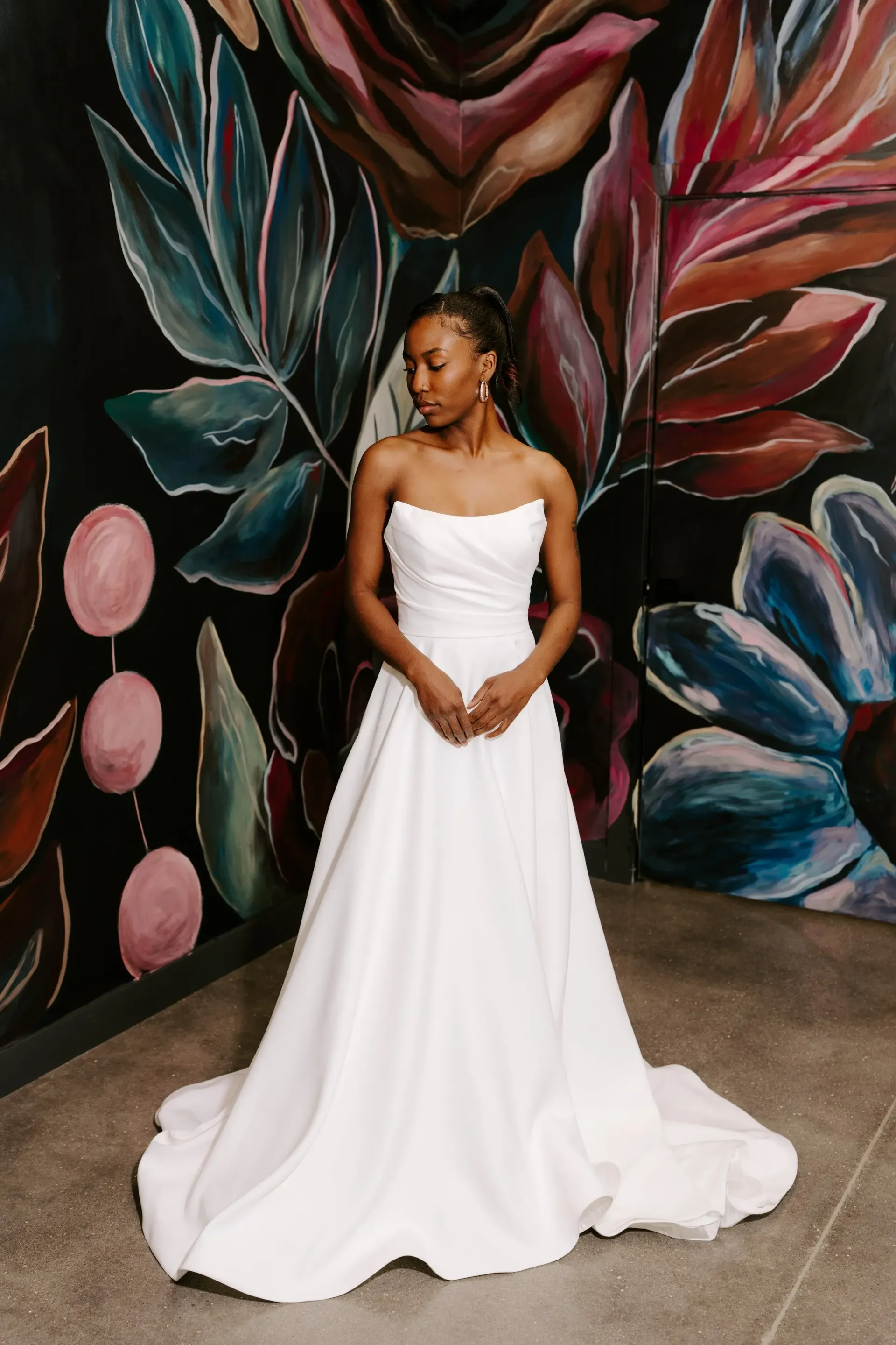 Woman in a white strapless wedding dress, standing in front of a colorful floral mural.