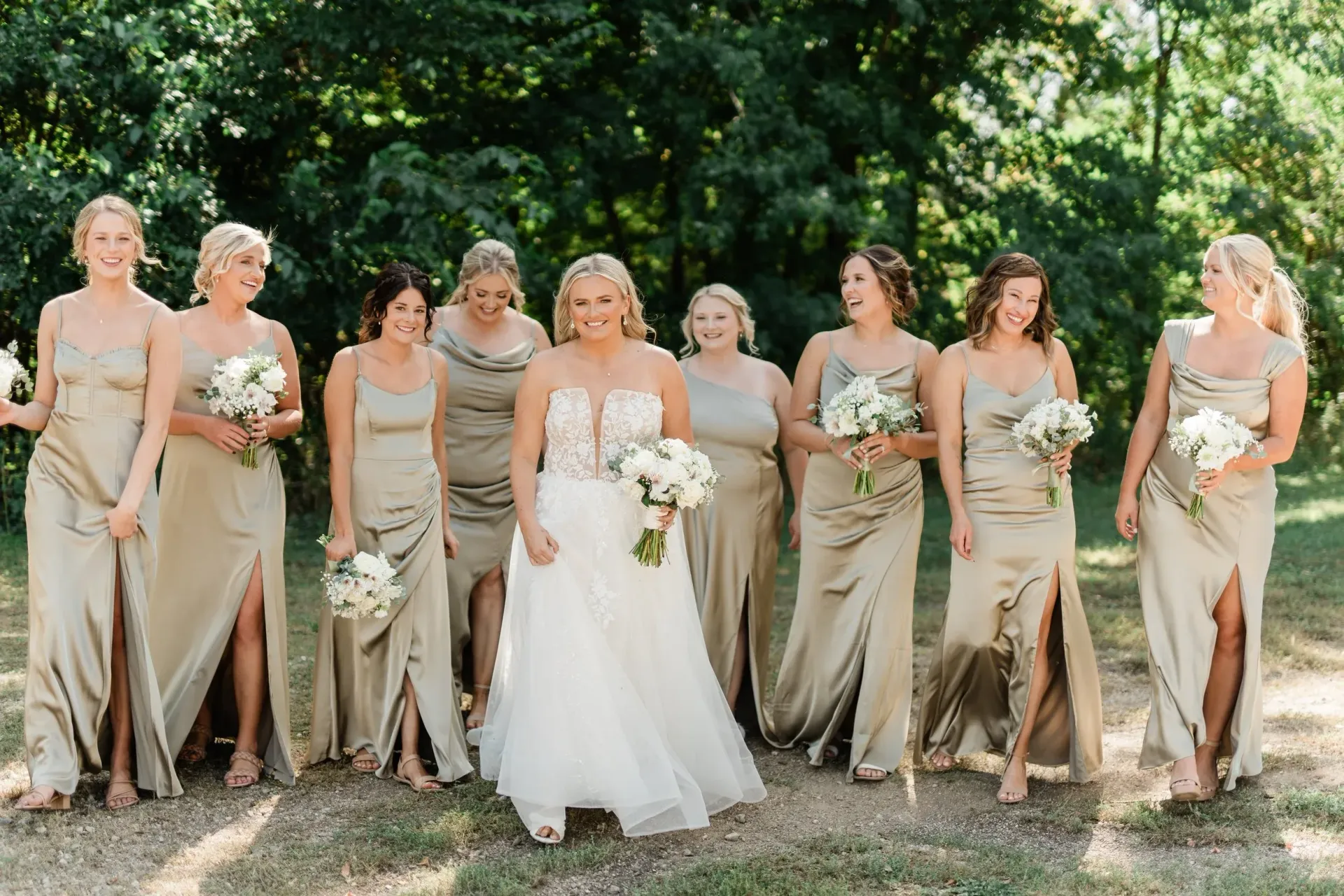 A bride in a white gown smiles with her bridesmaids, who wear gold dresses and hold bouquets, outdoors.