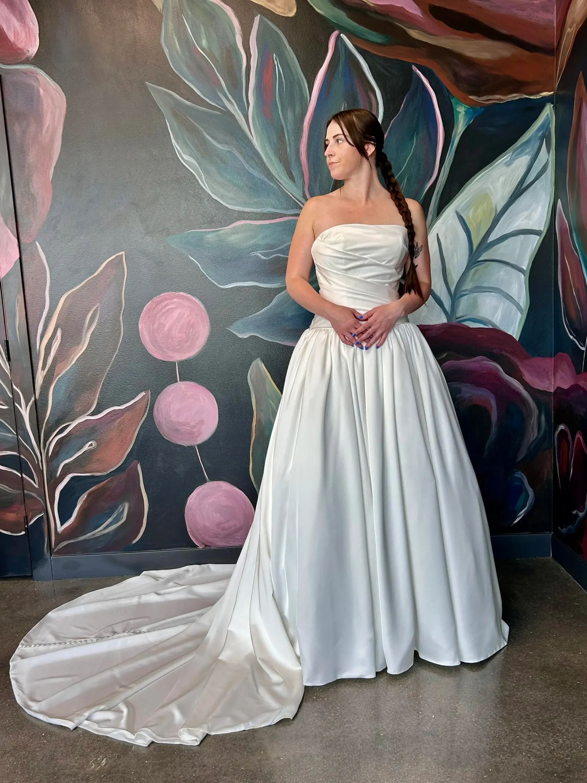 Woman in a white strapless wedding dress with a train, standing in front of a floral mural.