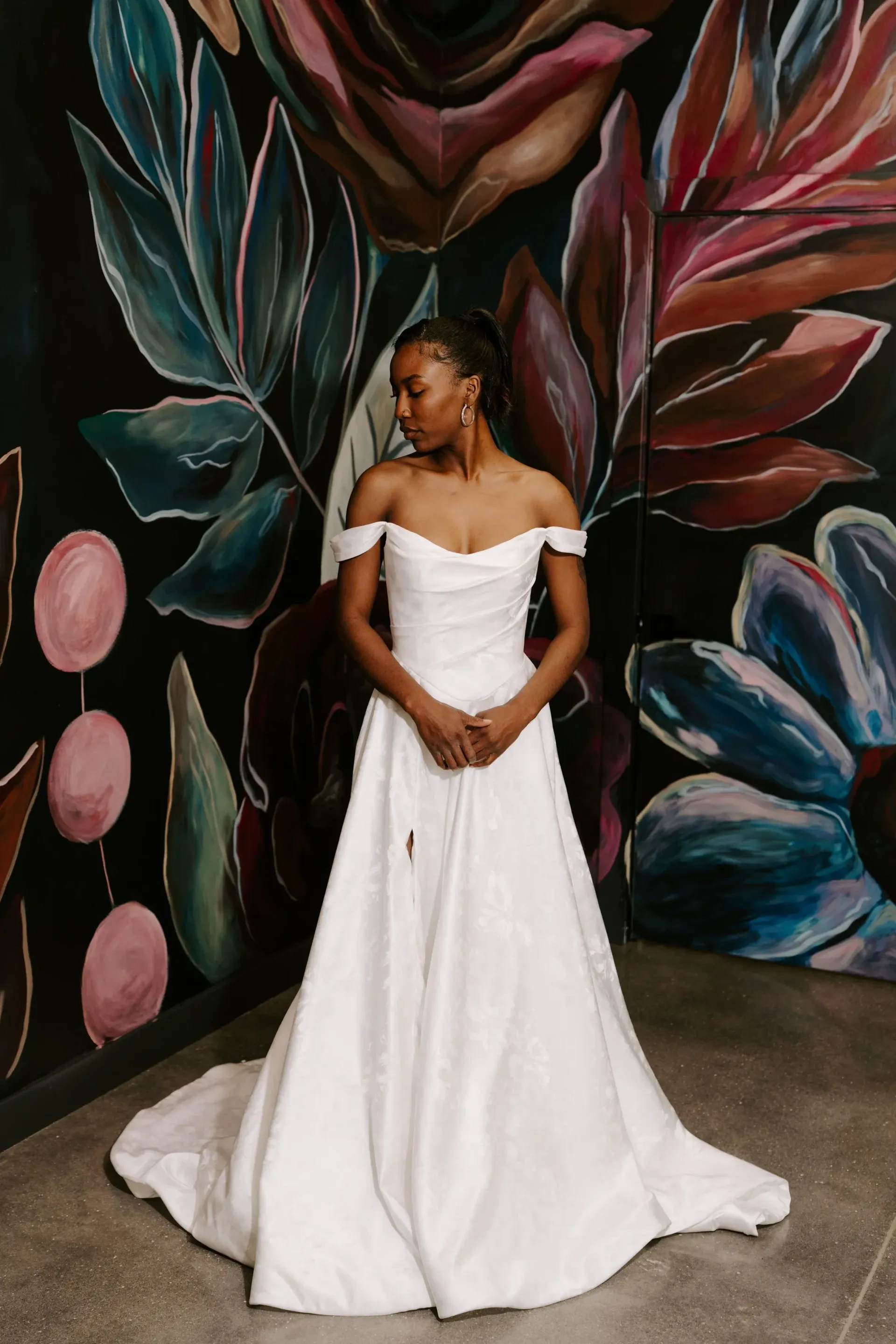 Bride in white off-the-shoulder gown, standing before colorful floral mural on black wall.