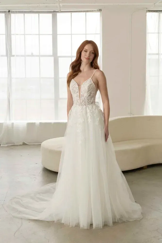 Woman in a white wedding dress, smiling, in front of a window and couch.