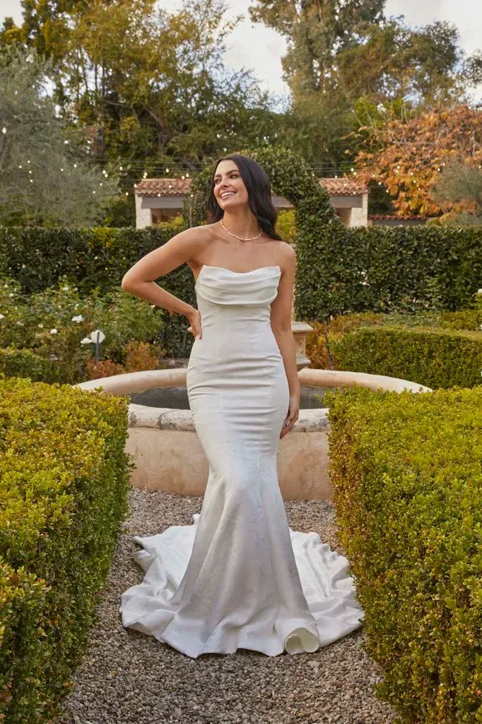 Woman in white strapless gown smiles in garden, posing by a fountain.