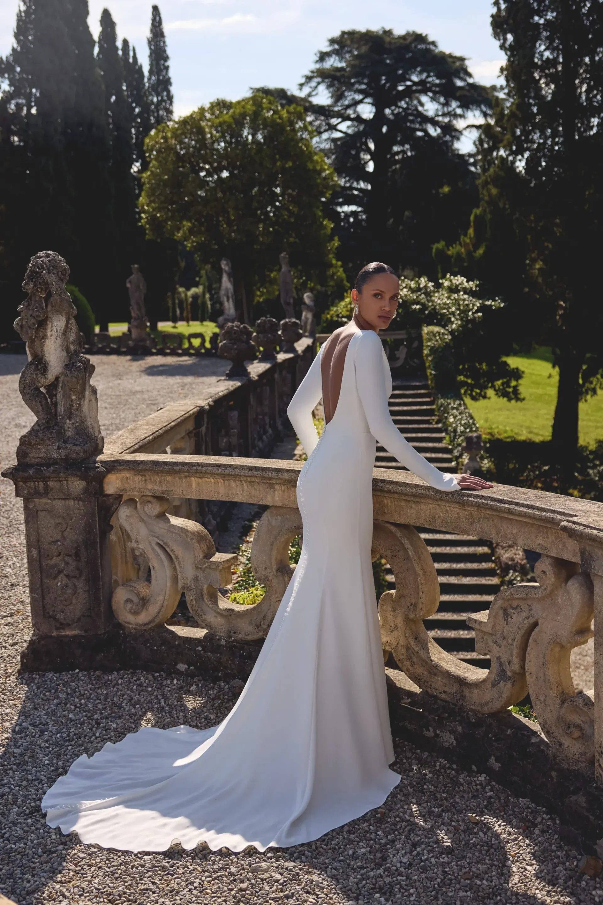 Woman in white gown stands on a stone balcony, looking back. Trees and ornate structures in the background.