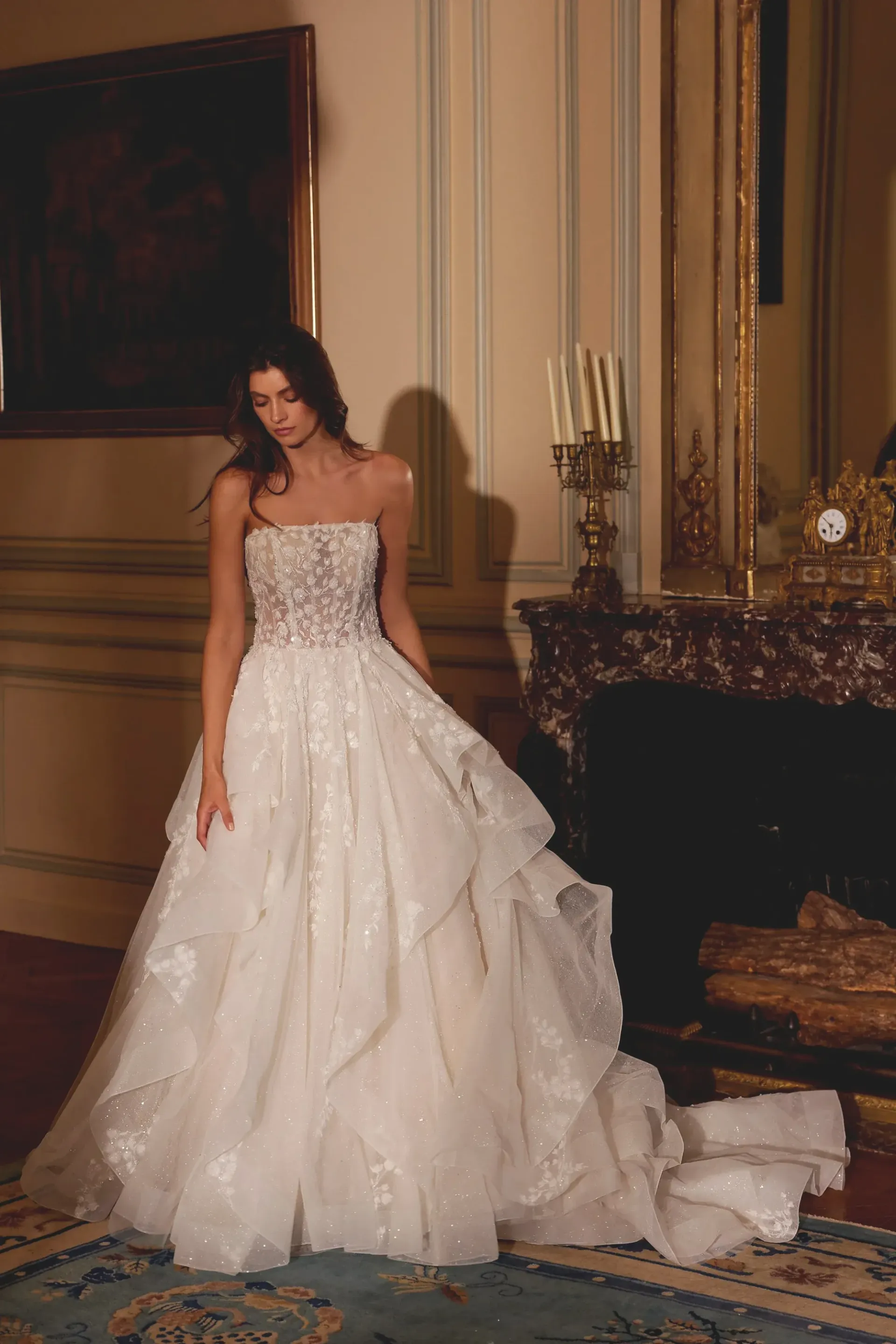 Woman in a white strapless ballgown stands in a room with a fireplace, looking down.