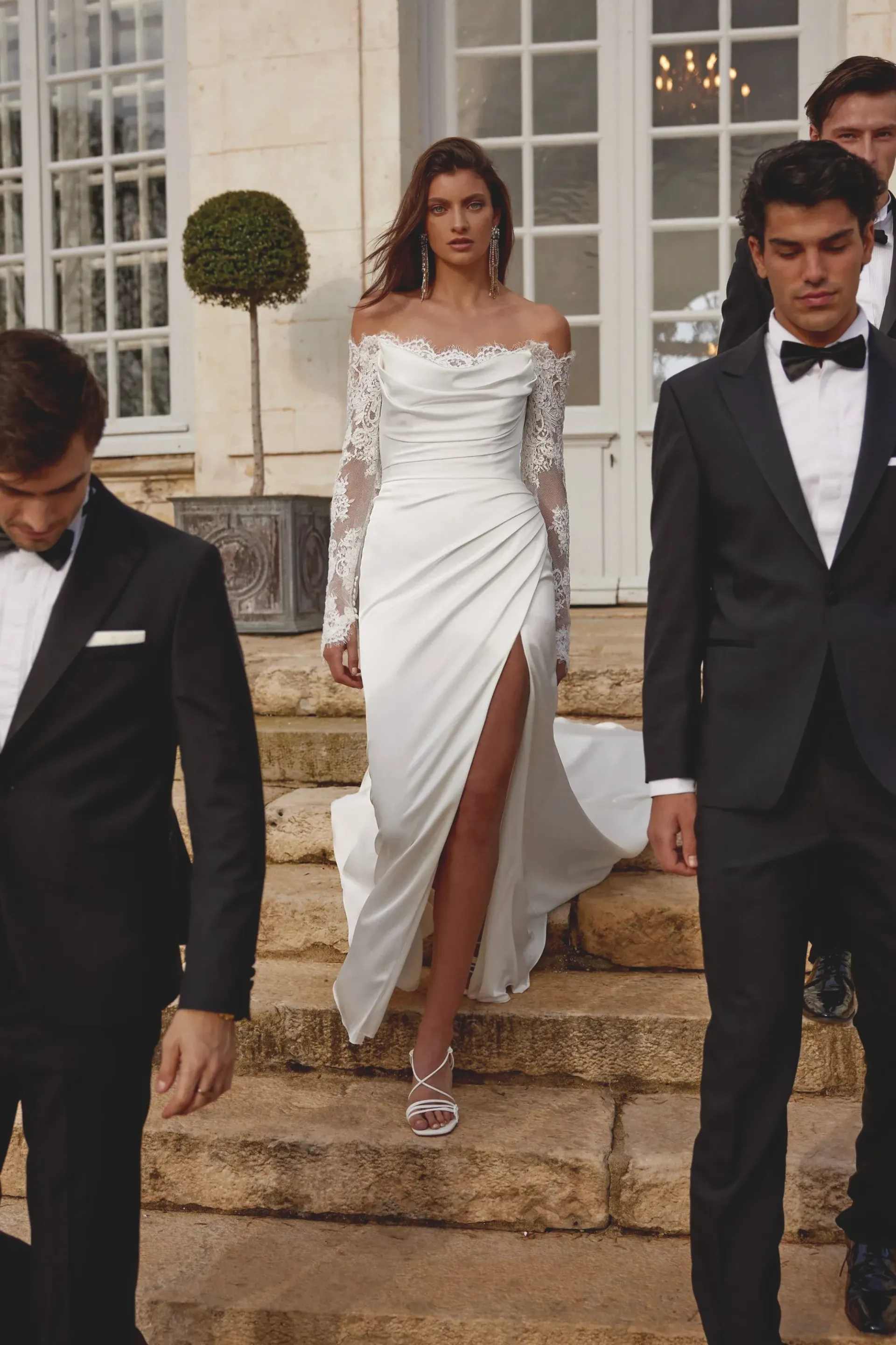 Woman in white gown with thigh-high slit descends stone steps, followed by men in black suits.