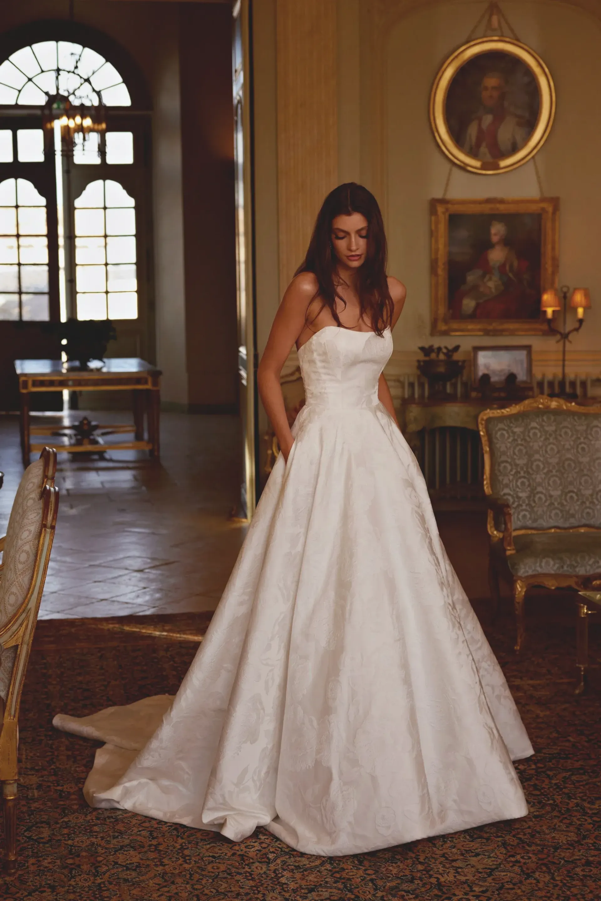 Woman in strapless wedding dress, hands in pockets, in a grand hall with paintings.