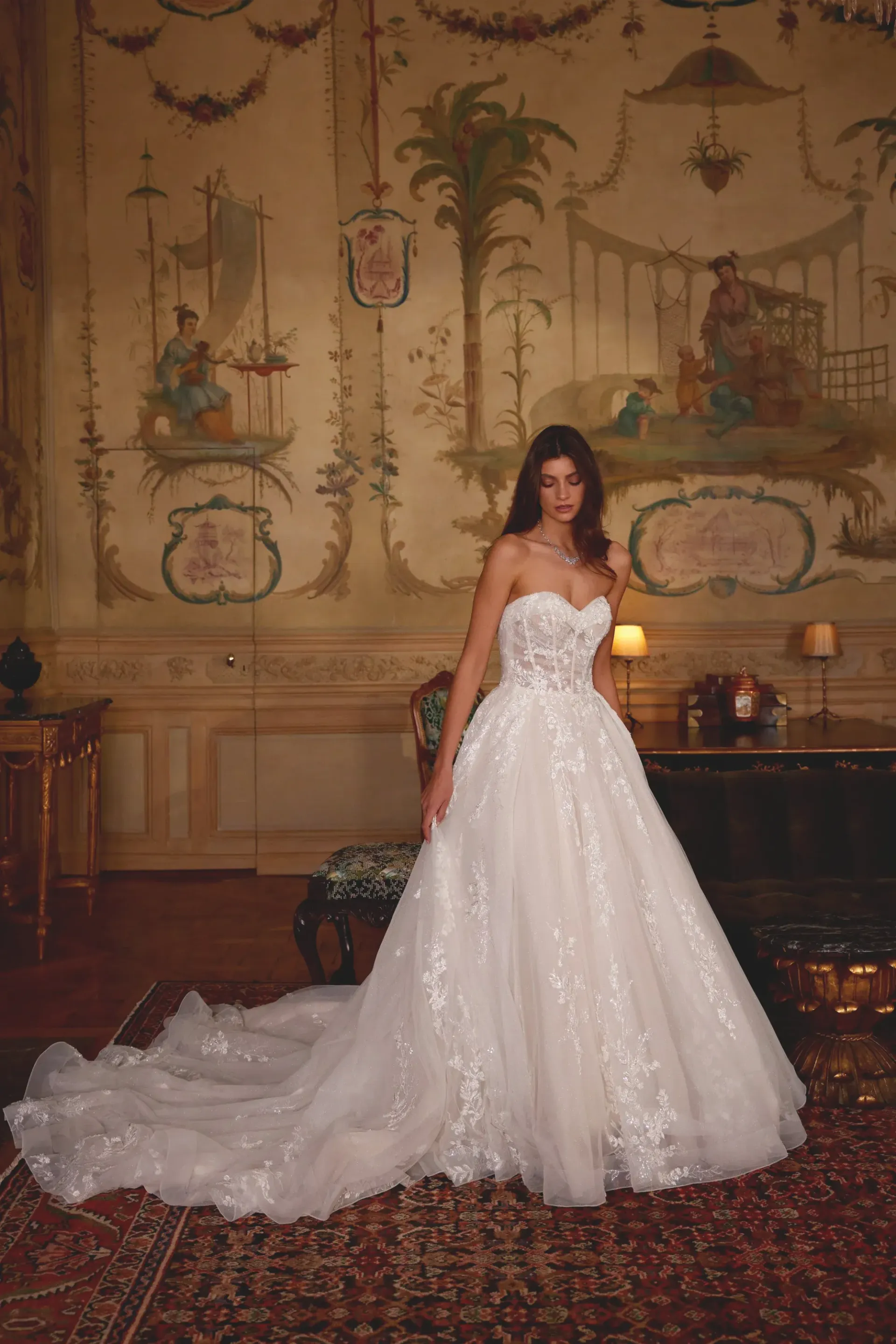 Woman in a white, strapless wedding gown, standing in an ornate room, train flowing behind her.