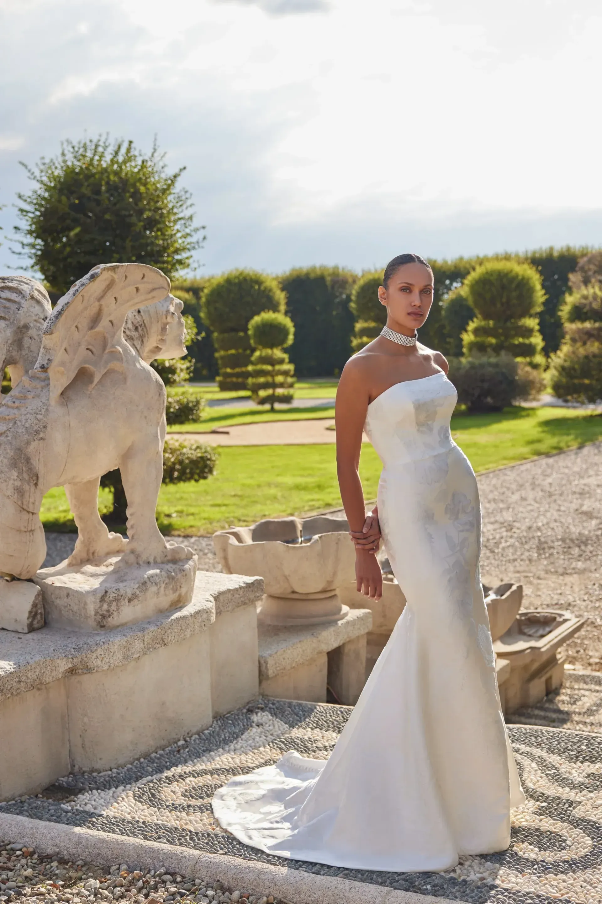 Woman in strapless white wedding dress poses by stone lion statue, garden background.