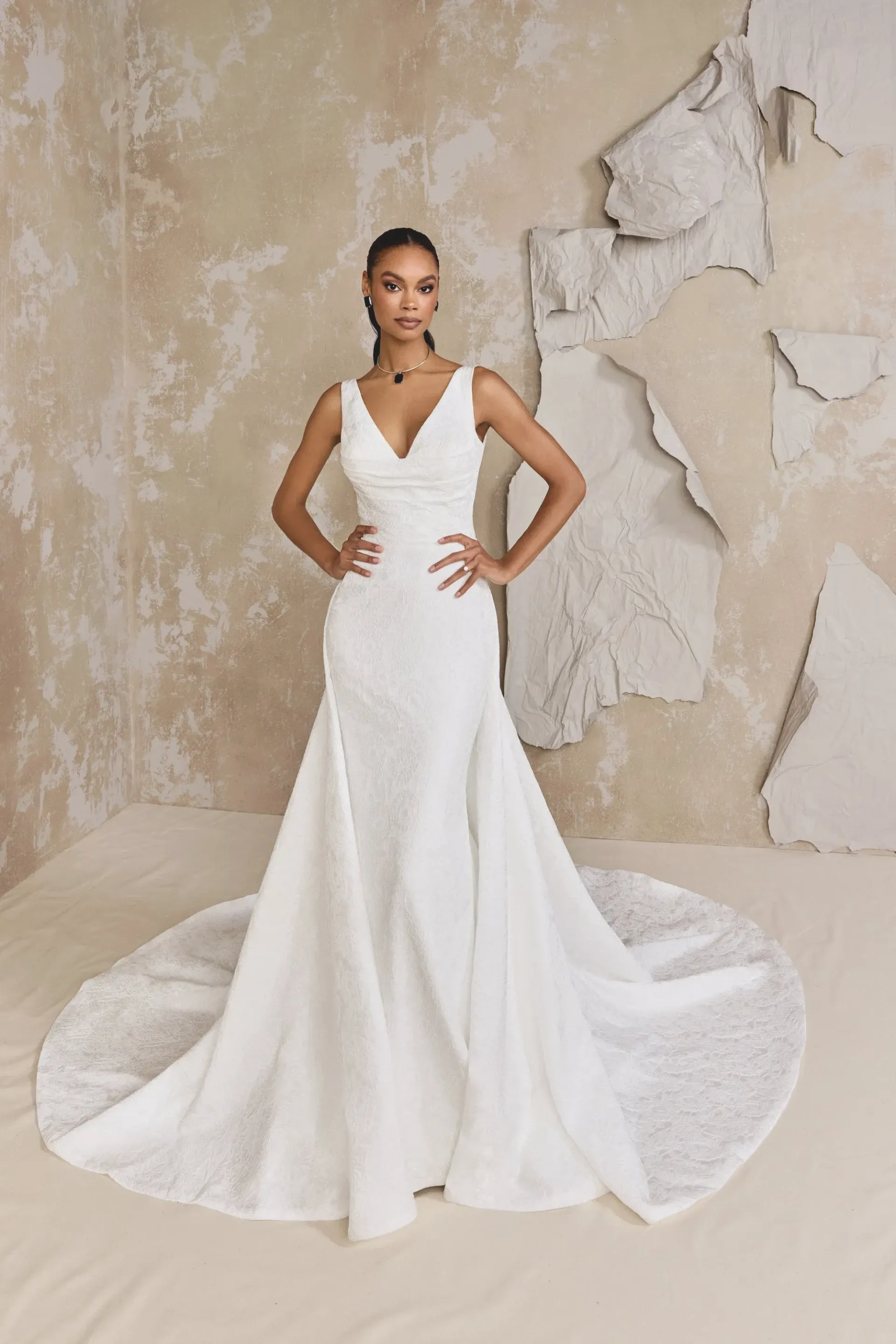 Woman in a white wedding dress with a V-neck and mermaid train, posing with hands on hips.