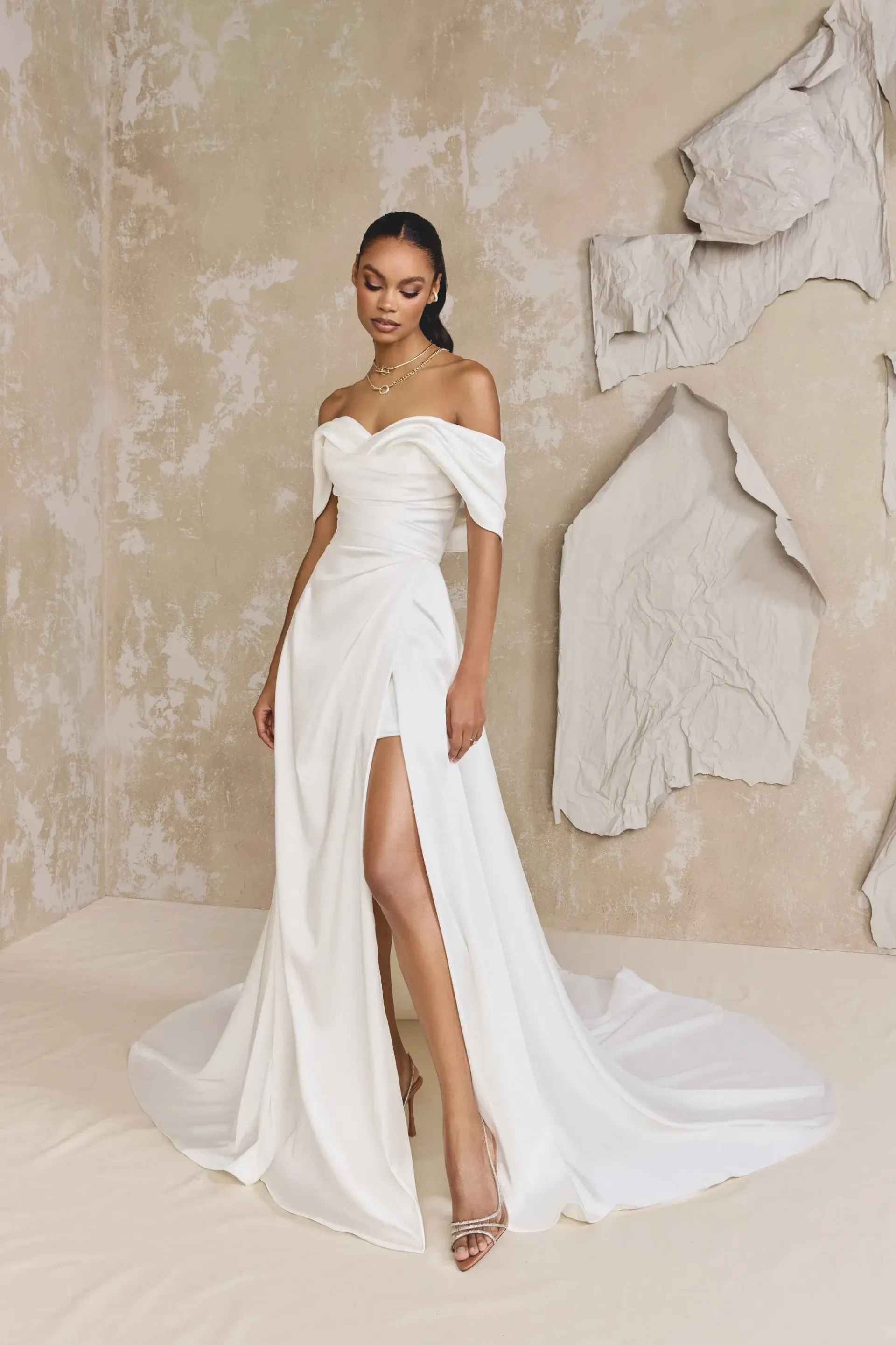 Woman in white off-shoulder wedding dress with high slit, standing in front of textured wall.