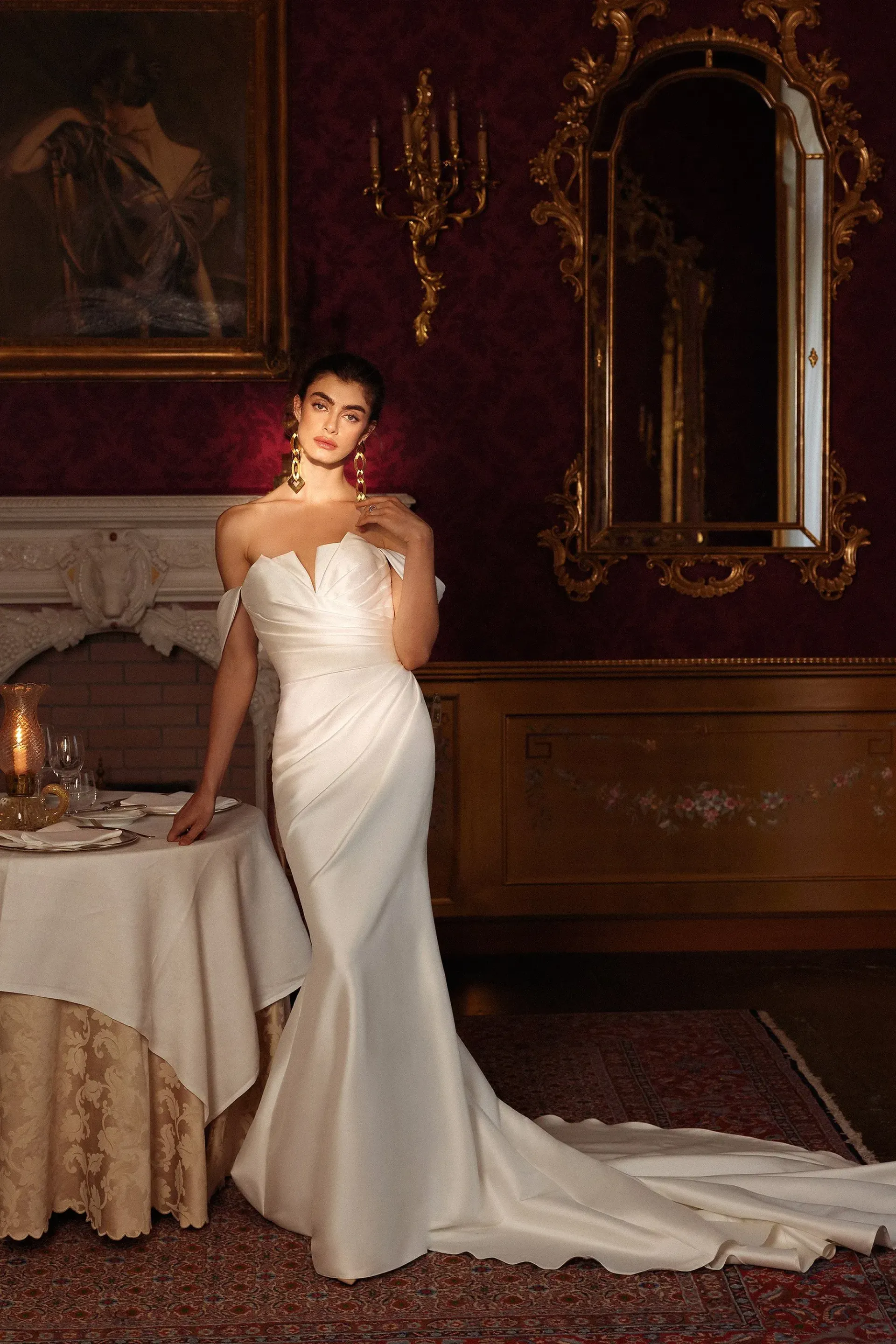 Woman in white gown poses elegantly by a table in a lavish room with antique decor.