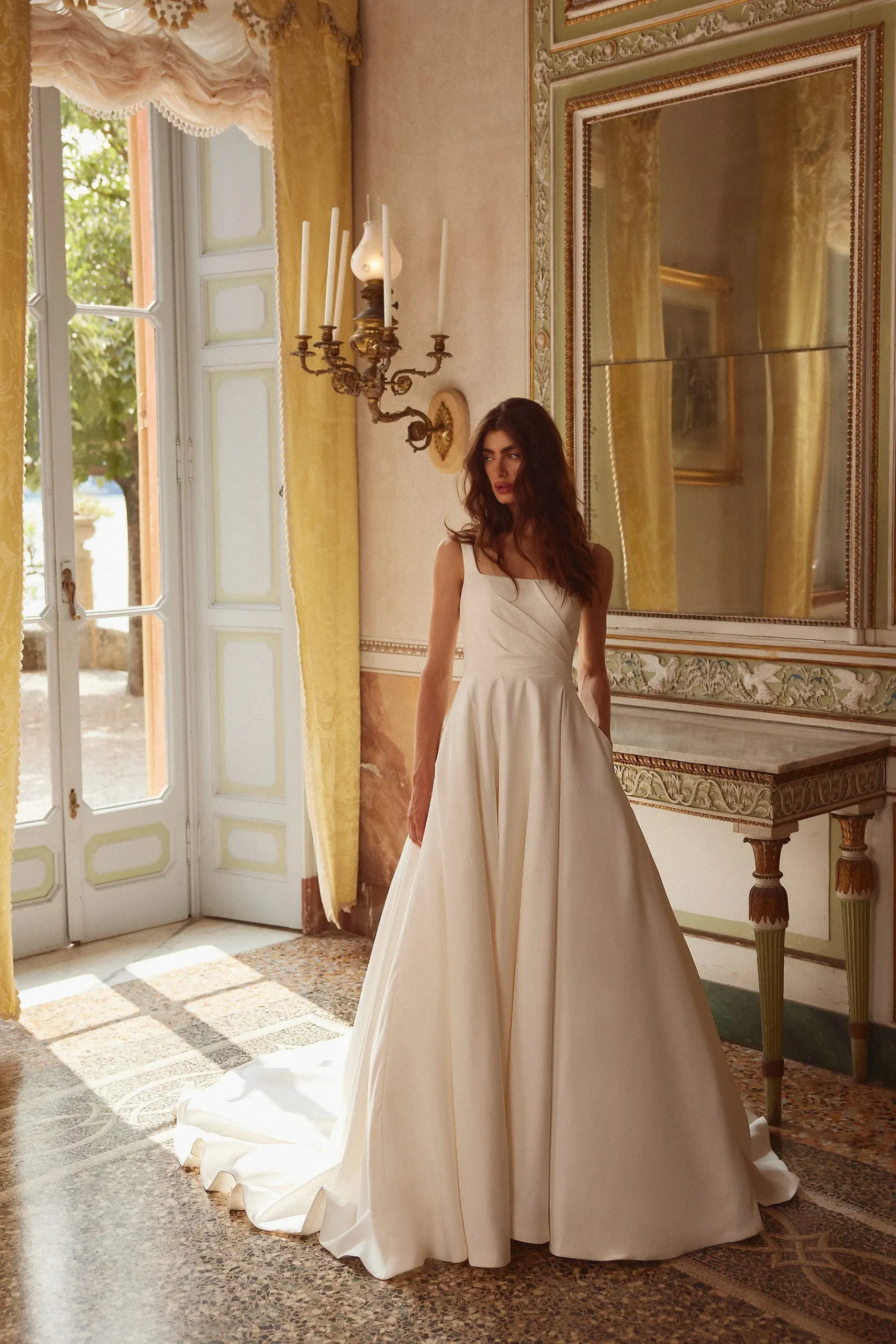 Woman in a white wedding dress stands in a sunlit ornate room.