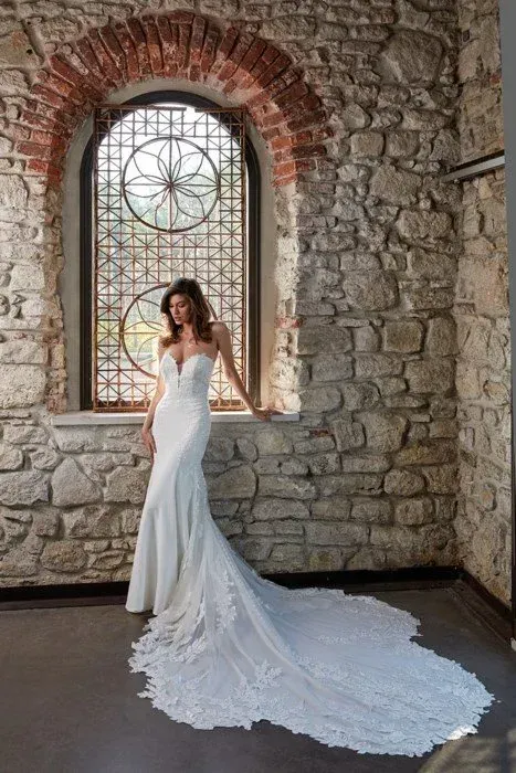 Bride in white strapless gown with long train, standing by arched window in stone building.