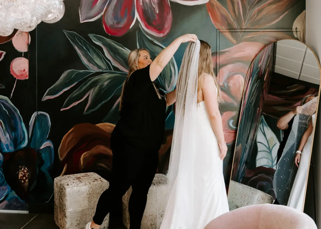 A woman in a white wedding dress has her veil adjusted by another woman in a floral painted room.
