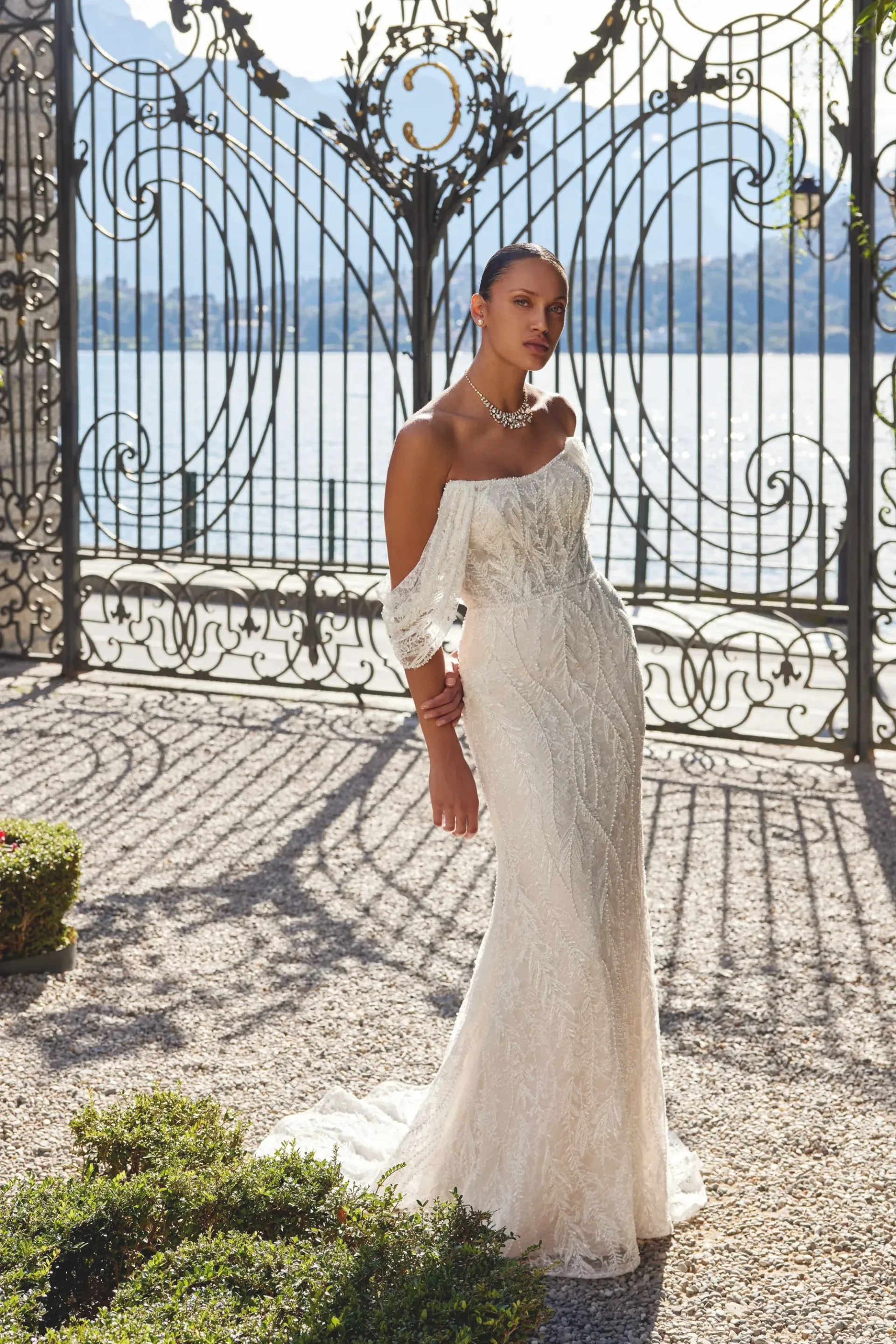 Woman in a beaded white gown stands before ornate gate, lake and mountains in the background.