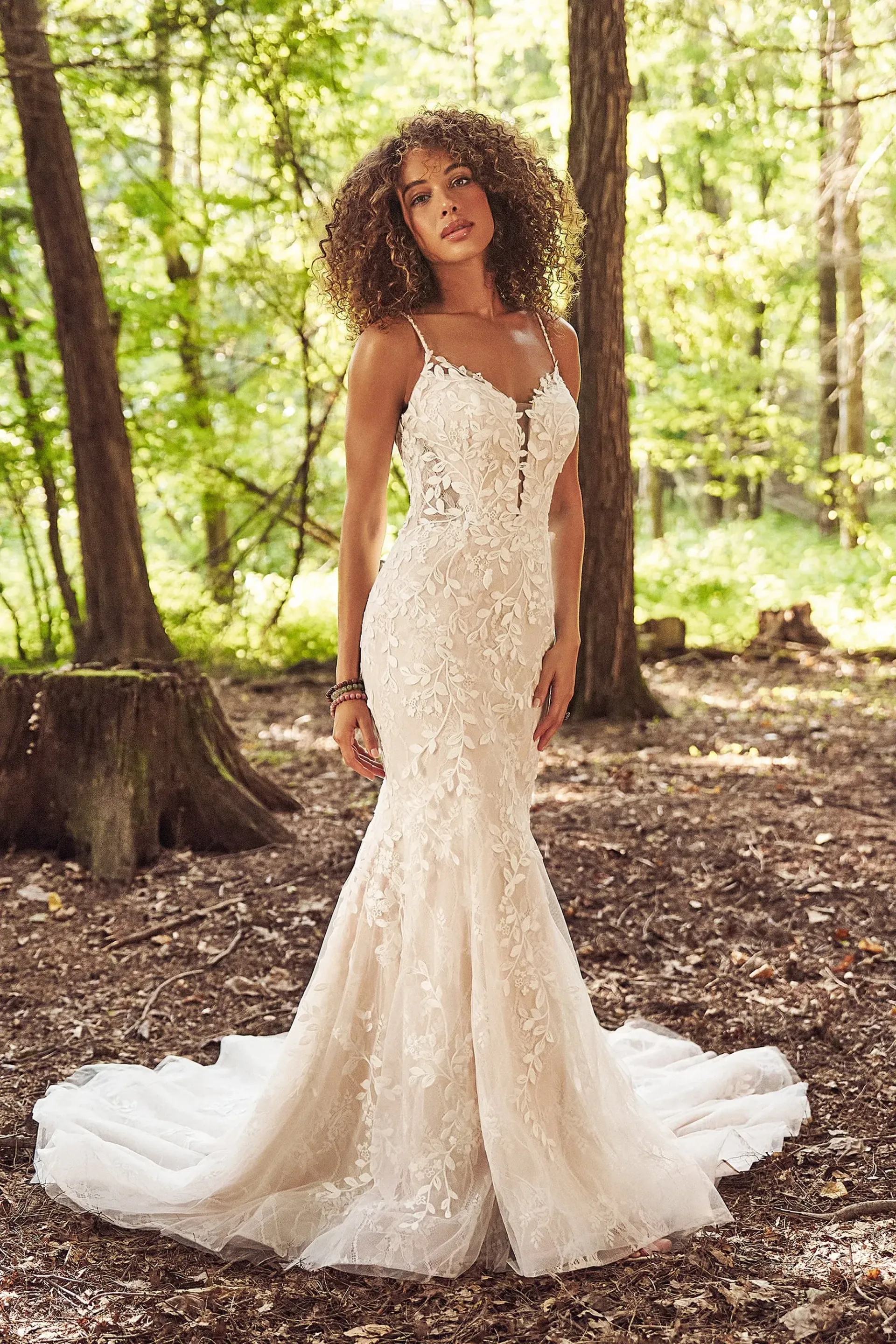 Woman in a white lace wedding dress with a train, standing in a forest.