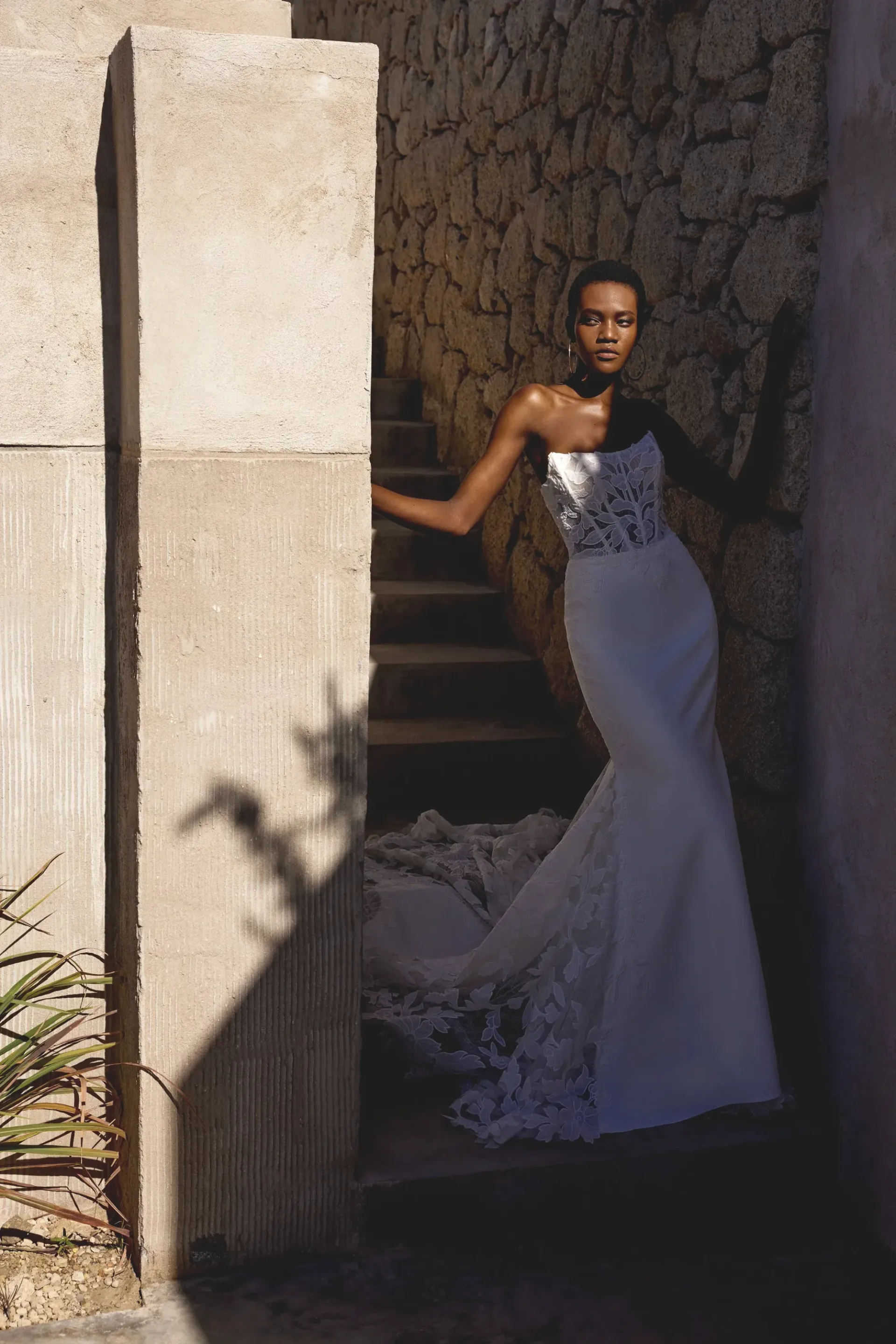 Woman in white wedding gown, standing on stairs in a sunlit stone alcove.