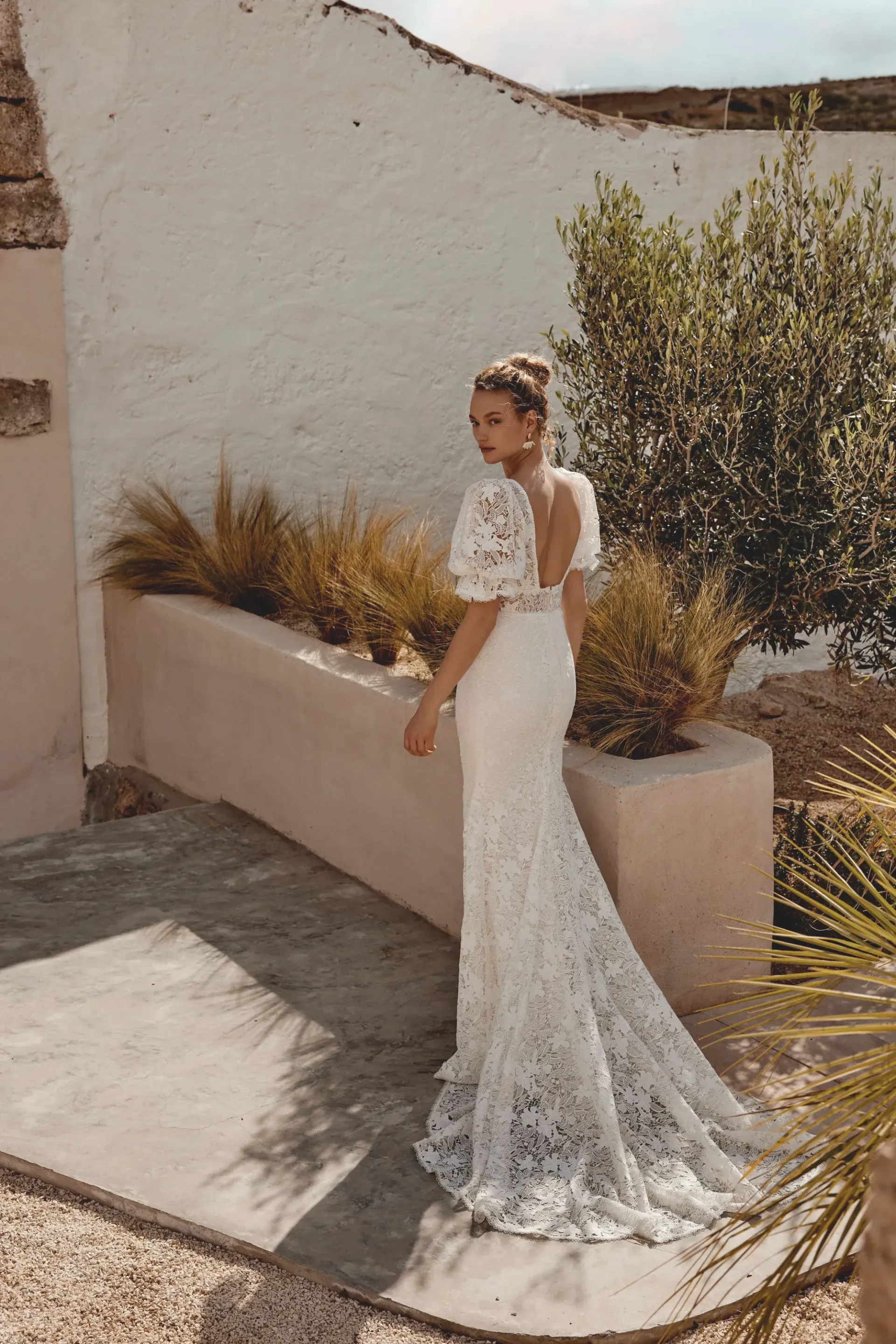 Woman in white lace wedding dress, looking over her shoulder. Outdoor setting, sunny.